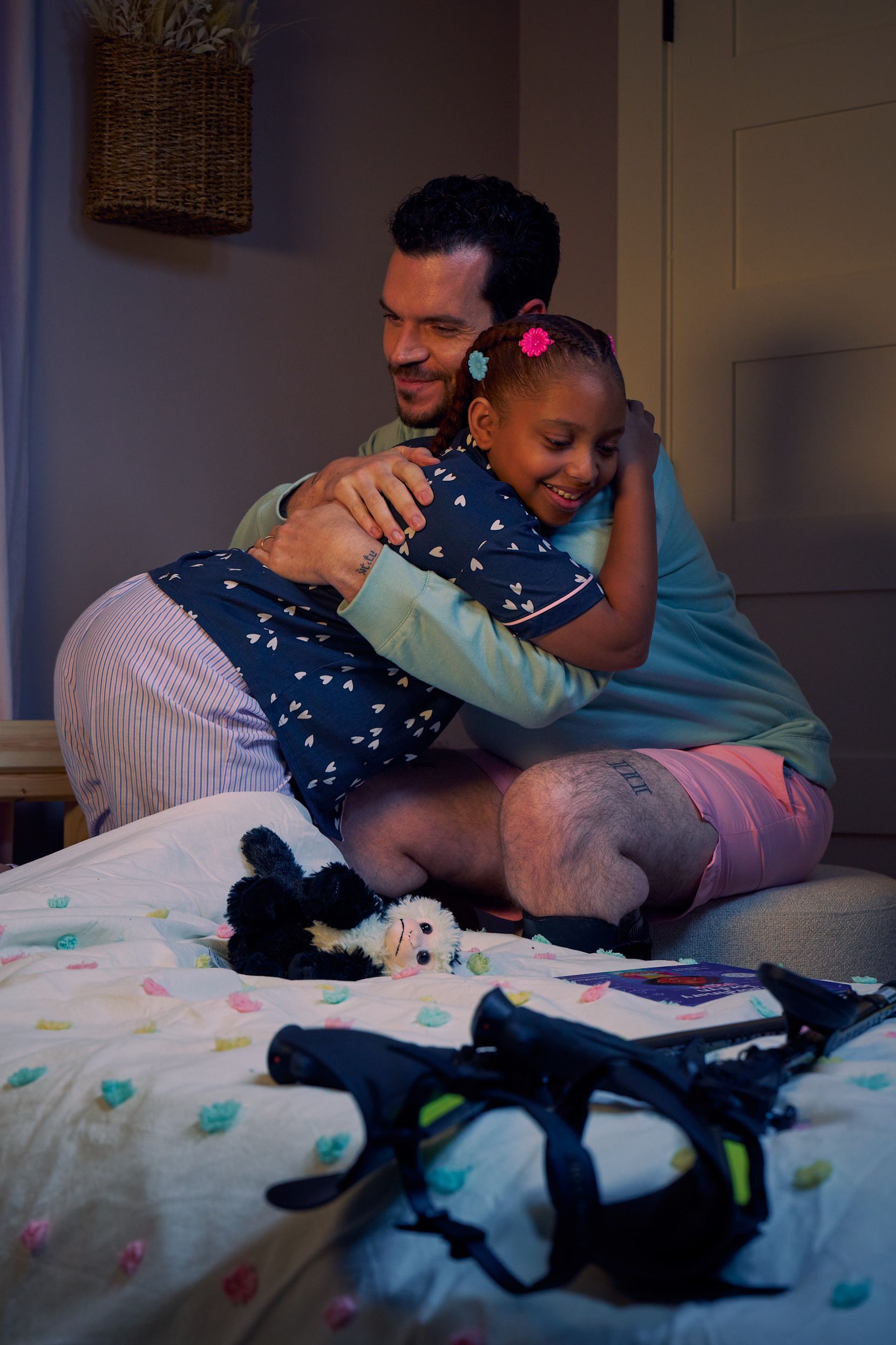 A father and daughter smile and embrace in her bedroom at night. The father is a white man with cerebral palsy who wears leg braces. His black forearm crutches lie on the foot of the daughter's bed. The daughter is a Black multiracial American girl with thick braided red hair adorned with two flower barrettes and is wearing blue patterned pajamas. A Capuchin monkey plushie lays on her bed.