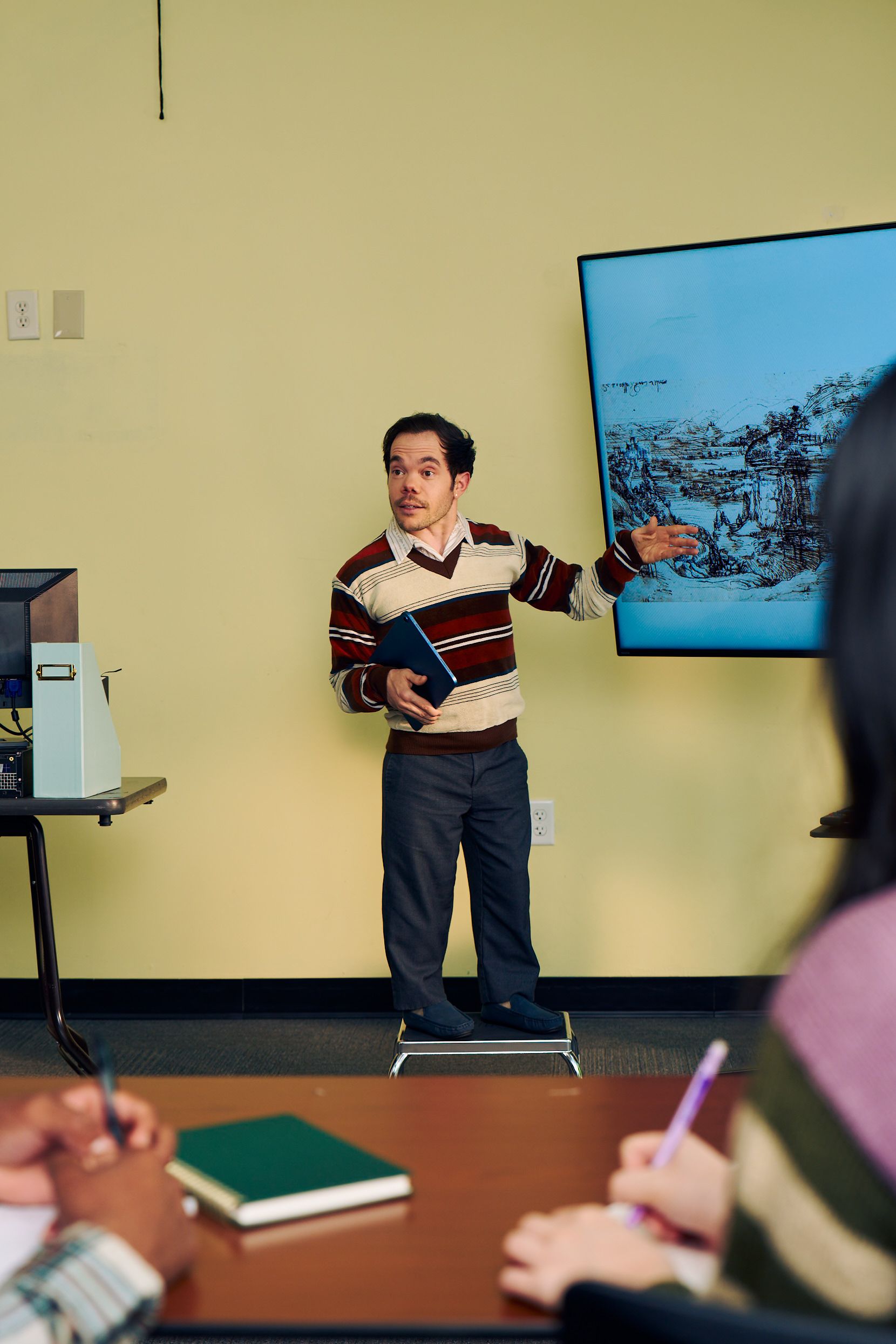 A white male professor with medium brown hair and light brown eyes who has dwarfism stands on a stool and gestures at a large monitor next to him, displaying a pen and ink drawing by Leonardo da Vinci. He is holding a tablet in his other hand. He has short brown and is wearing a red striped sweater, a collared shirt, and dark dress pants. In front of him, two students write in notebooks.