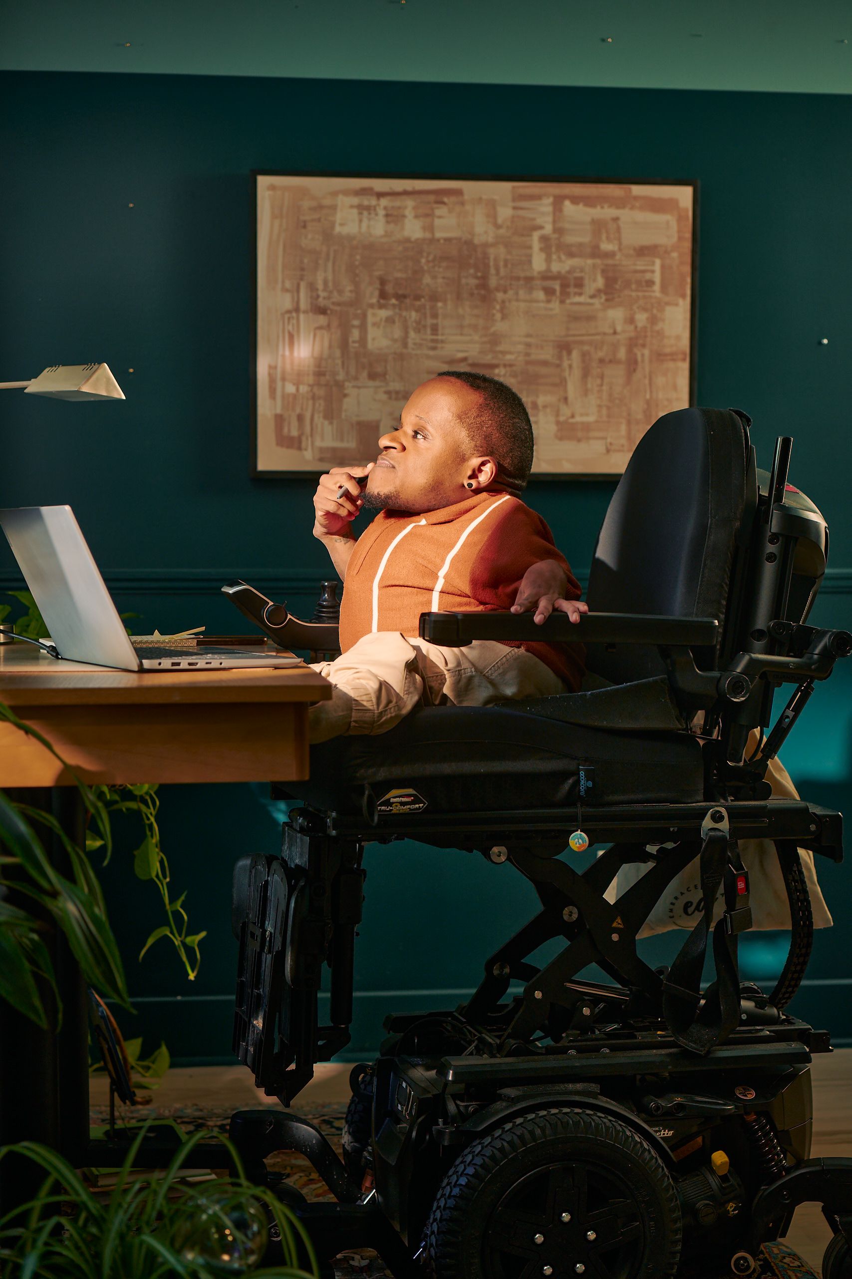 A black man who is a little person is seated in his power wheelchair in a dark teal home office. He holds a pen in his hand up to his chin and looks away, deep in thought. A laptop, notebook, pen, and lamp are on the desk in front of him. His wheelchair is in an elevated position, allowing him to be level with his desk. His desk is surrounded by several house plants and an abstract art piece. He has closely cropped black hair, brown eyes, and is wearing a rust-orange knit quarter-zip sweater with white vertical stripes, light khaki trousers, and a small stud earring. A tote bag hangs on the back of his wheelchair.