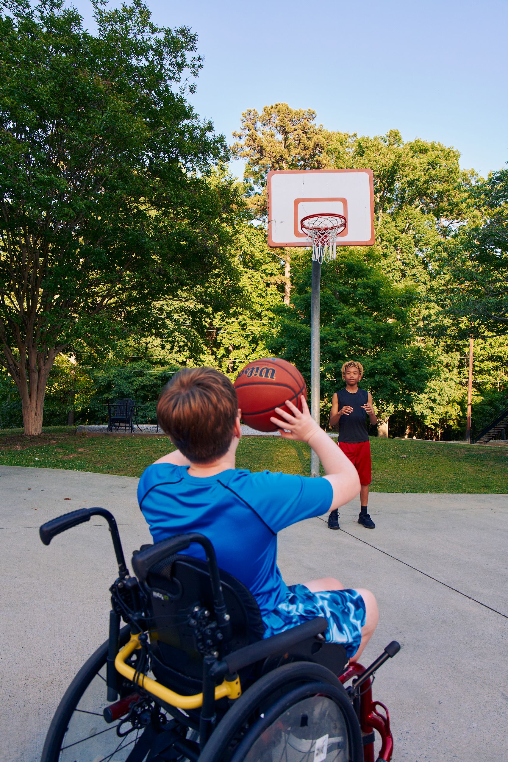 Two teens play basketball at an outdoor park. One of the friends, a white boy seated in a manual wheelchair, passes the ball to the other, a light-skinned African American male, who is standing near the hoop. The teen seated in the manual wheelchair has short brown hair is wearing blue athletic wear and faces away from the camera. The African American teen has curly brown to blonde hair and is wearing black and blue athletic wear.