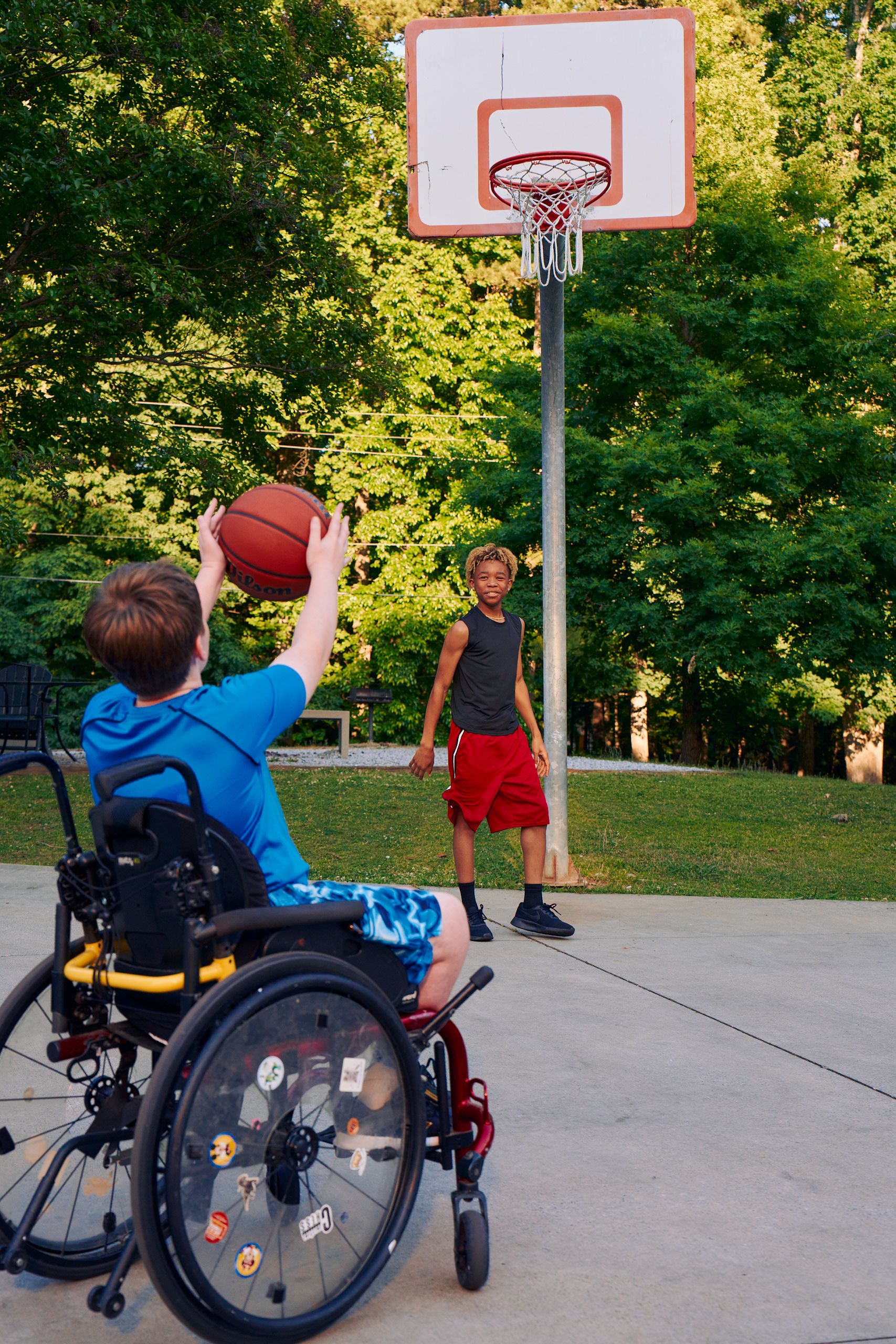 Two teens play basketball at a neighborhood park. One of the friends, a white boy seated in a manual wheelchair, passes the ball while a light-skinned African American male, who is standing near the hoop, watches. The teen seated in the manual wheelchair has short brown hair is wearing blue athletic wear and faces away from the camera. The African American teen has curly brown to blonde hair and is wearing black and blue athletic wear.