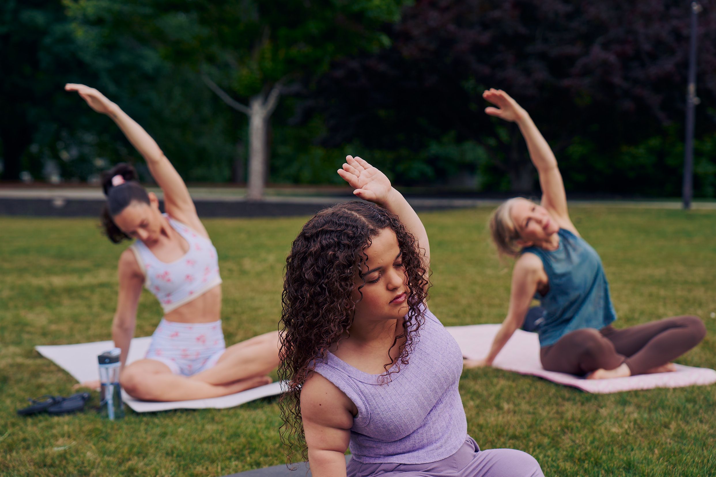 At an outdoor yoga class, a young white woman with dwarfism sits in side bend stretch, cross-legged with her eyes closed, one hand resting on the ground while the opposite arm reaches up and arches over to the side. She has long, curly brown hair and is wearing a purple ribbed tank with comfortable, loose purple pants. Behind her, two tall women sit on their mats in the same pose. The class is in a grassy park with many large trees.