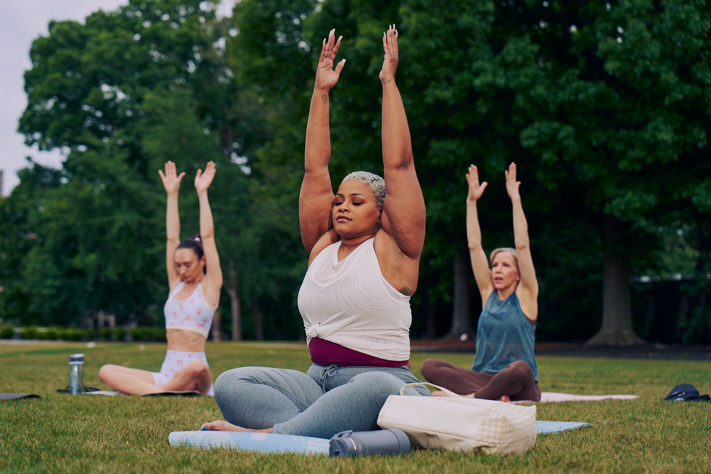 At an outdoor yoga class, a Black woman sits cross-legged with her eyes closed and hands extended straight overhead with palms facing inward toward each other. Her bag and water bottle are next to her. She has icy blonde shortly cut hair and is wearing a white and red layered tank and gray drawstring pants. Behind her, two tall women sit on their mats in the same pose. The class is in a grassy park with many large trees.