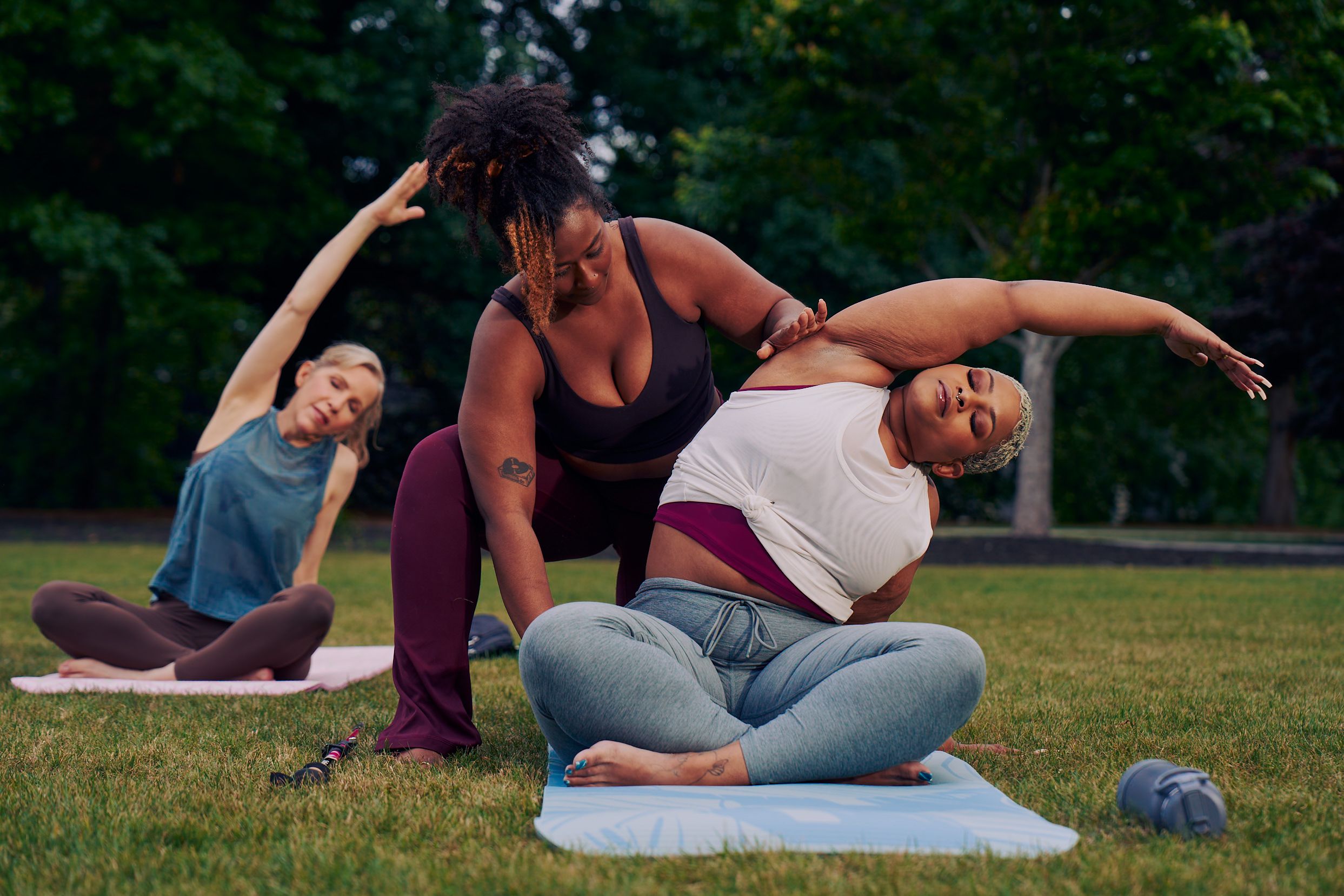 A Black woman yoga instructor supports a Black woman student move into a modified side bend stretch. The student sits in a cross-legged position, leaning to her right side with her left arm reaching over to stretch her left side. The instructor stands behind her, gently placing one hand on her outstretched arm, and the other on her upper thigh. The student has a walking cane placed on the grass next to her yoga mat. She has icy blonde shortly cut hair and is wearing a white and red layered tank and gray drawstring pants. The instructor has her black hair pulled back into a natural ponytail with dyed bangs and is wearing a black tank and maroon yoga pants. Behind her, another woman sits in the same pose, unsupported.