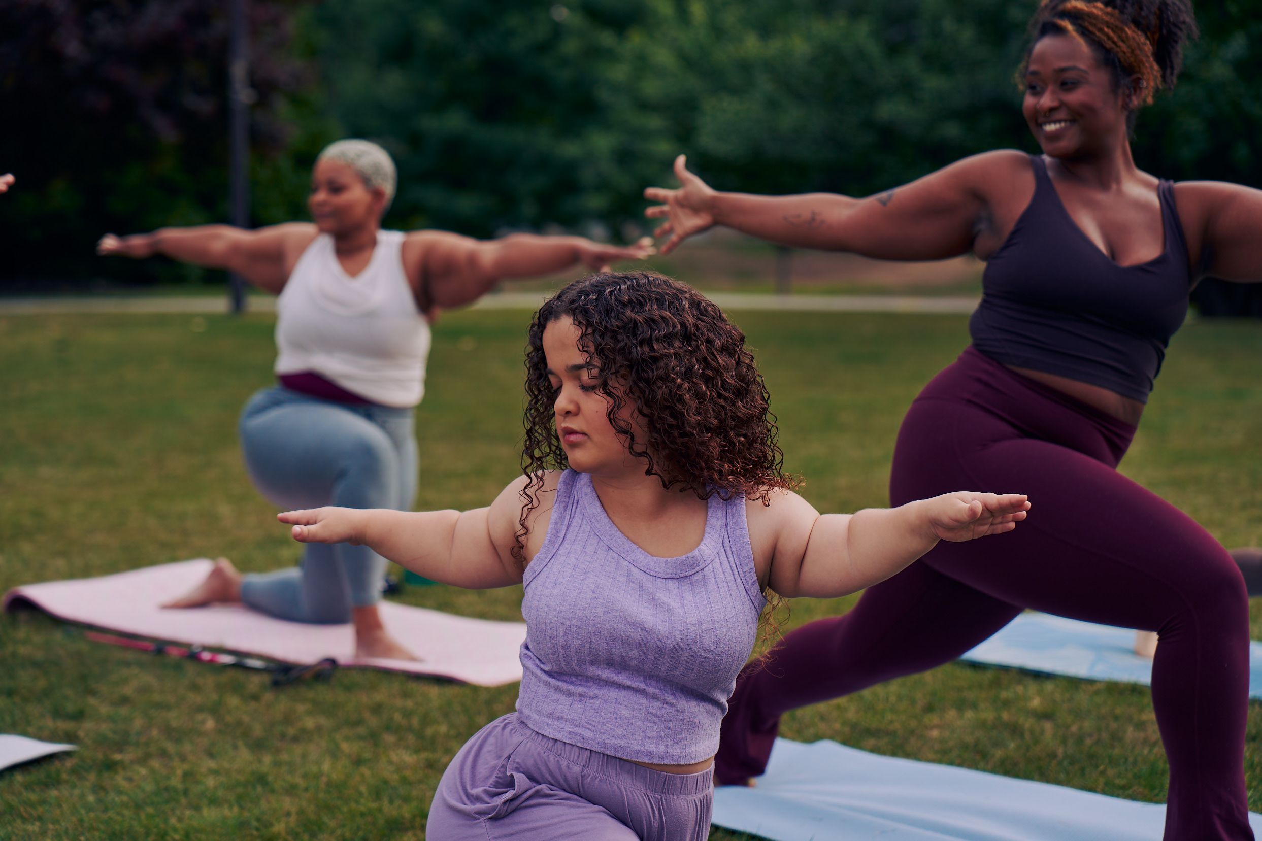 At an outdoor yoga class, a young white woman with dwarfism stands in Warrior pose, with her eyes closed, both arms extended out parallel to the ground, one facing forward and one reaching back. She has long, curly brown hair and is wearing a purple ribbed tank with comfortable, loose purple pants. Behind her, a tall Black woman instructor smiles while also in the same pose. Another Black woman is in a modified form of the pose, kneeling. Her cane is next to her mat. The class is in a grassy park with many large trees.