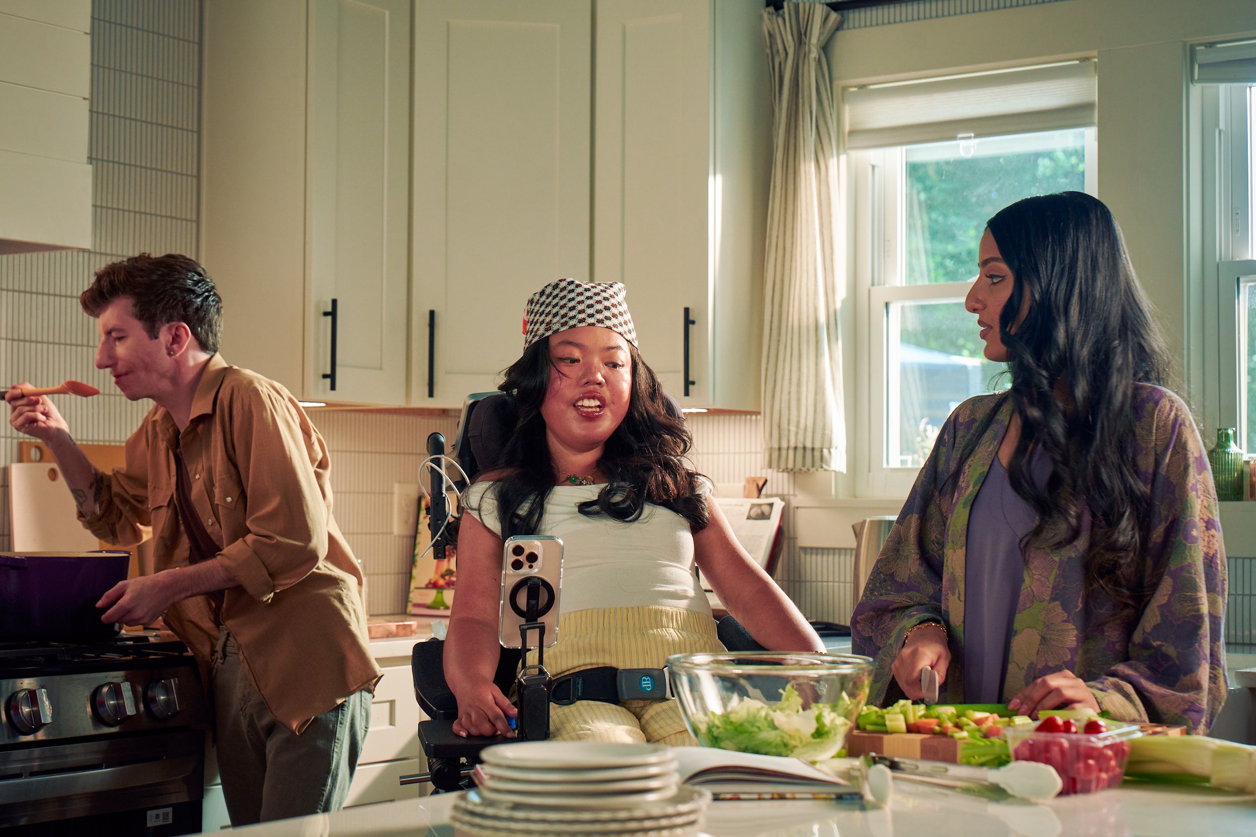 A Southeast Asian woman with curled black hair seated in her power wheelchair gives her roommate instructions on how to prepare a salad in their bright kitchen. Her smart phone is mounted to her wheelchair. Behind them, a white male with Moebius Syndrome and a face that is paralyzed holds a wooden spoon up to his lips to taste a soup on the stove. The Southeast Asian woman is wearing a white top with striped, yellow linen pants and a checkered headscarf. He has short, wavy, reddish brown hair and is wearing a tan button down shirt with the sleeves rolled up, showing arm tattoos. Their roommate has long dark hair and is wearing a purple floral dress.