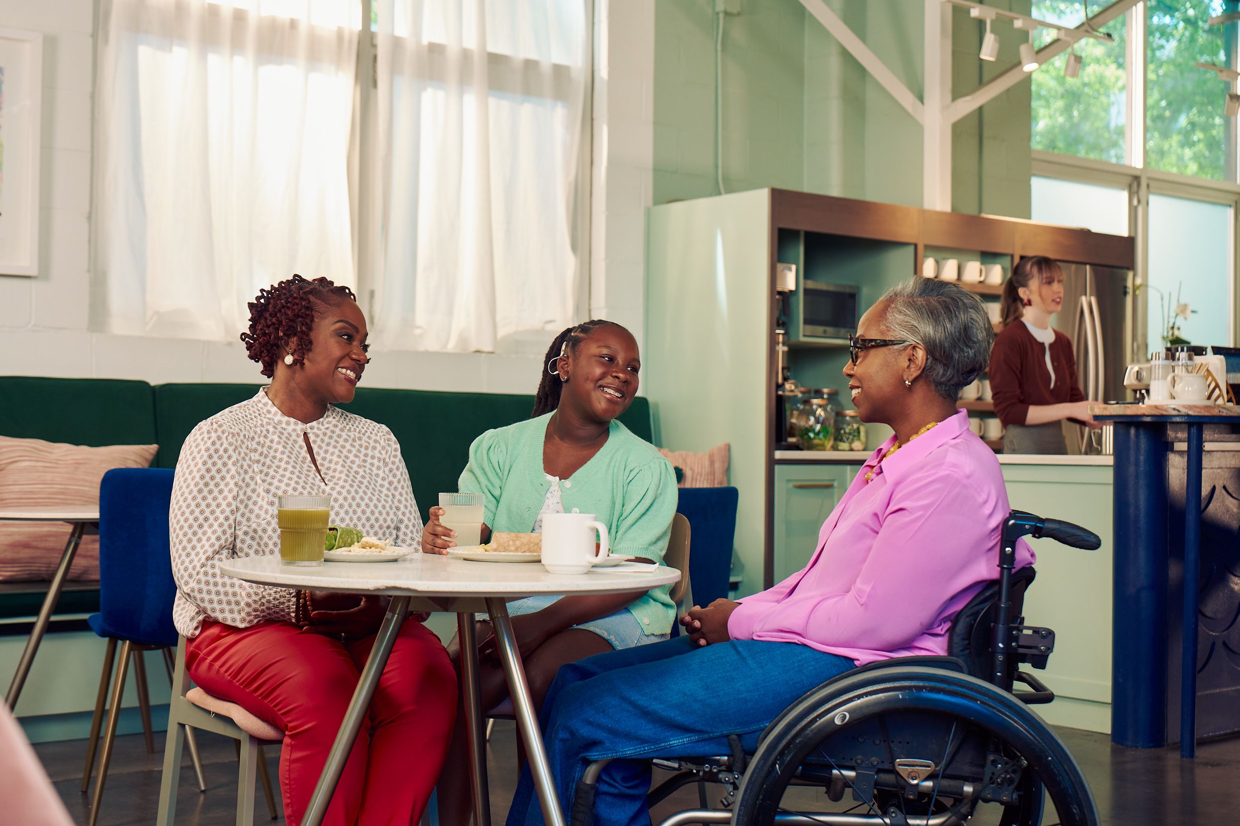 An African American mother, daughter, and auntie are smiling while chatting at a table in a modern cafe. The auntie is paraplegic and is seated in a manual wheelchair. She has short gray hair is wearing a lavender silk blouse and dark blue jeans. The mother has short tightly curled red hair and is wearing a patterned blouse and red pants. The daughter is wearing a cochlear implant and has her braided hair pulled back into a pony tail. She is wearing a light green cardigan and jean shorts. Behind them, a barista prepares drinks and a man types on a laptop.