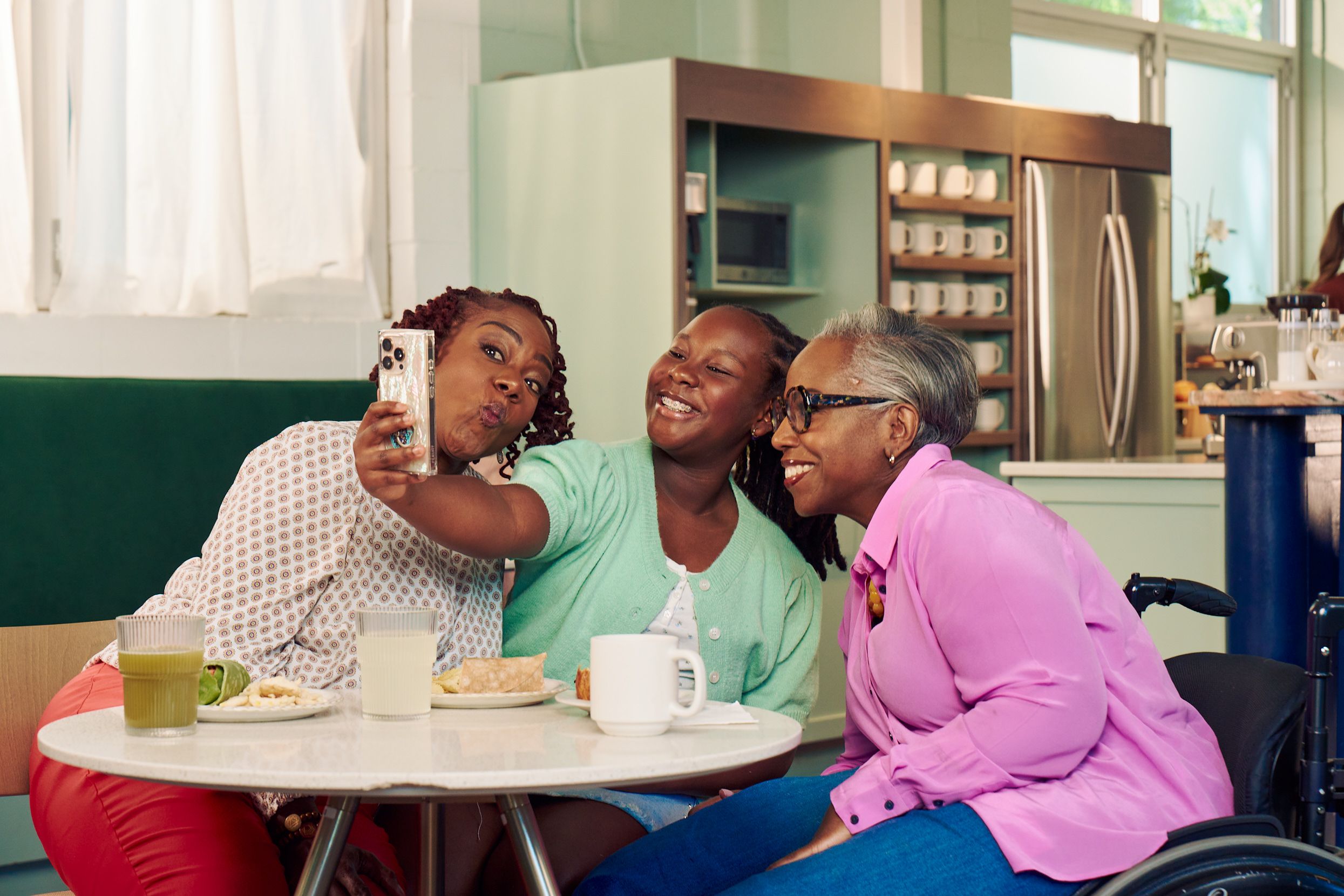 An African American girl takes a selfie with her mother and auntie in a modern cafe. The auntie is paraplegic and is seated in a manual wheelchair. She has short gray hair is wearing a lavender silk blouse and dark blue jeans. The mother has short tightly curled red hair and is puckering her lips in a silly pose. She is wearing a patterned blouse and red pants. The daughter is wearing a cochlear implant and has her braided hair pulled back into a pony tail. She is wearing a light green cardigan and jean shorts.