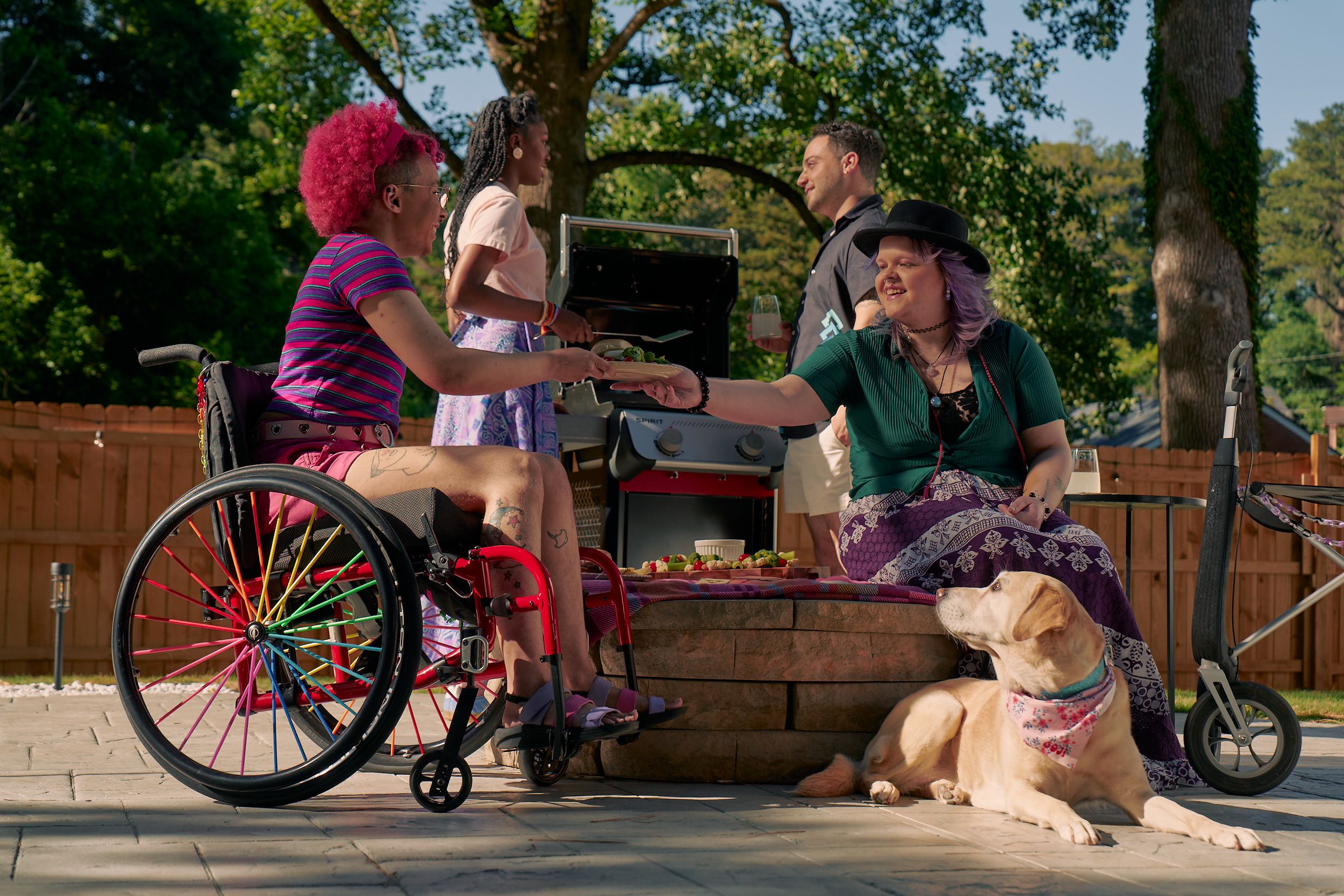 At a backyard barbeque, a white nonbinary person sits on a low stone table and passes a plate of food to a Black nonbinary person seated in a manual wheelchair with rainbow spokes. The white nonbinary person has a uniquely shapped dark gray rollator decorated with purple acrylic chains nearby. They have layered purple hair with two thin long braids on each side and are wearing a black hat, a green shirt, and a purple patterned skirt. The Black nonbinary person seated in their manual wheelchair has a curly pink fro with loc bangs and is wearing a purple and pink striped crop top, pink shorts, and purple sandals. They have several tattoos on both legs. A golden labrador retriever wearing a pink hankerchief looks up at the plate of food. Behind them, two friends chat at a grill. One friend is an African American woman and is holding a spatula. The other friend is a multi-ethnic man with curly black hair.