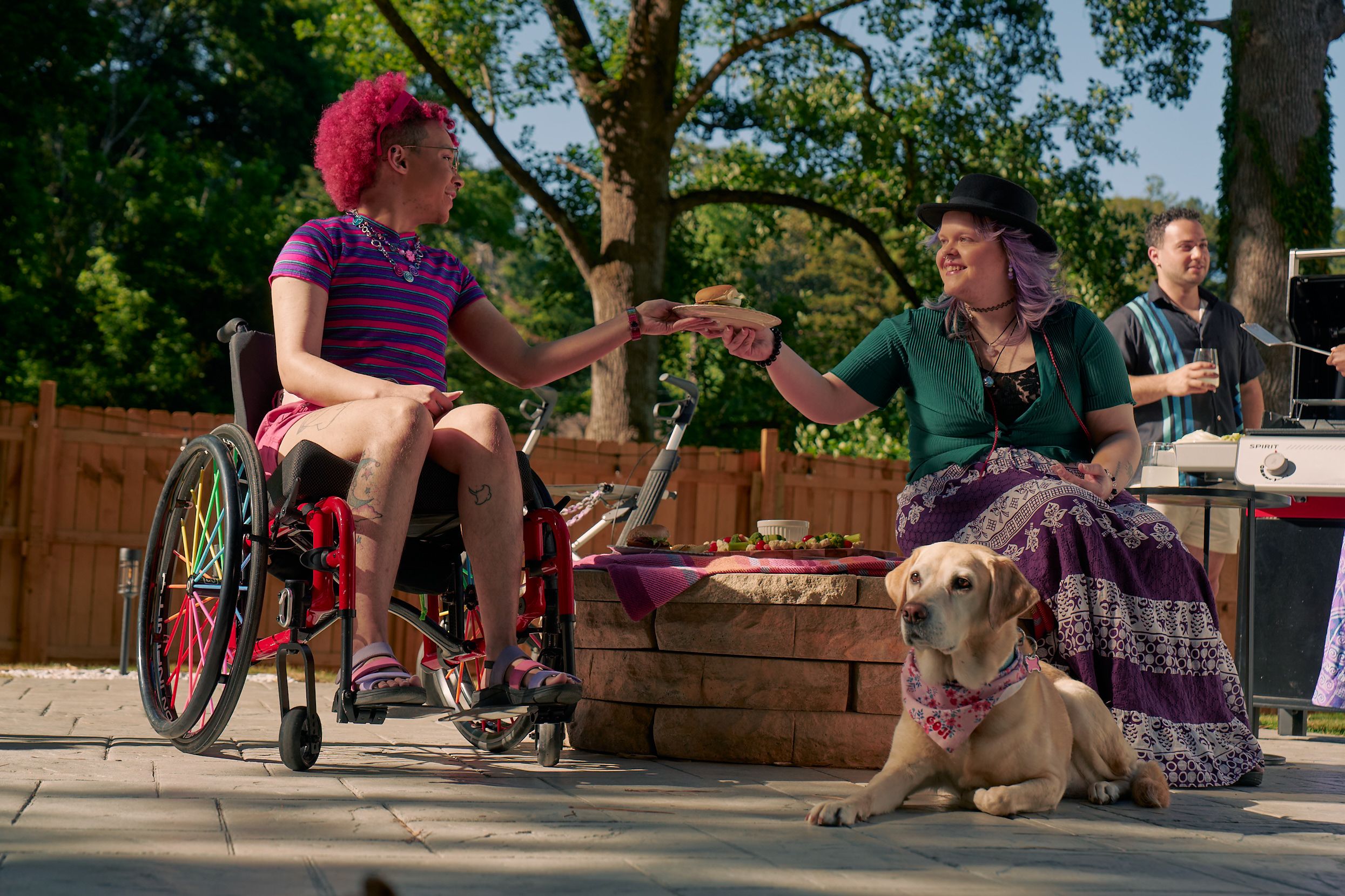 At a backyard barbeque, a white nonbinary person sits on a low stone table and passes a plate of food to a Black nonbinary person seated in a manual wheelchair with rainbow spokes. The white nonbinary person has a uniquely shapped dark gray rollator decorated with purple acrylic chains nearby. They have layered purple hair with two thin long braids on each side and are wearing a black hat, a green shirt, and a purple patterned skirt. The Black nonbinary person seated in their manual wheelchair has a curly pink fro with loc bangs and is wearing a purple and pink striped crop top, pink shorts, and purple sandals. They have several tattoos on both legs. A golden labrador retriever wearing a pink hankerchief sits at their feet. Behind them, another friend, a multi-ethnic man with curly black hair, holds a drink and chats near a grill.