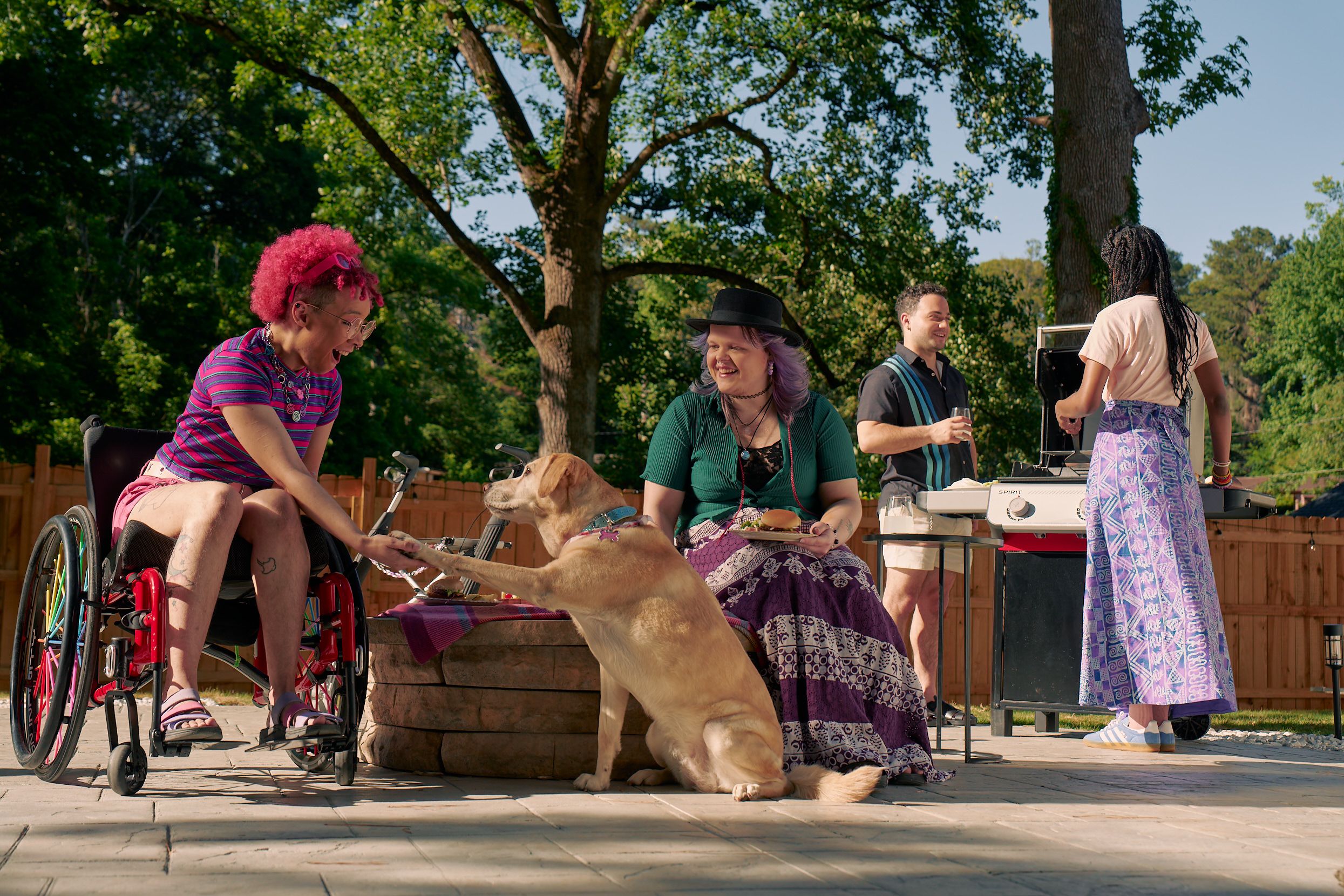 At a backyard barbeque, a golden labrador retriever places her paw in the palm of a Black nonbinary person's hand. The Black nonbinary person is seated in a manual wheelchair with rainbow spokes and has their mouth open in praise of the dog. Next to them, a white nonbinary person sits on a low stone table and holds a plate of food. They have layered purple hair with two thin long braids on each side and are wearing a black hat, a green shirt, and a purple patterned skirt. The Black nonbinary person seated in their manual wheelchair has a curly pink fro with loc bangs and is wearing a purple and pink striped crop top, pink shorts, and purple sandals. They have several tattoos on both legs. Behind them, two friends chat at a grill. One friend is an African American woman and is holding a spatula. The other friend is a multi-ethnic man with curly black hair.