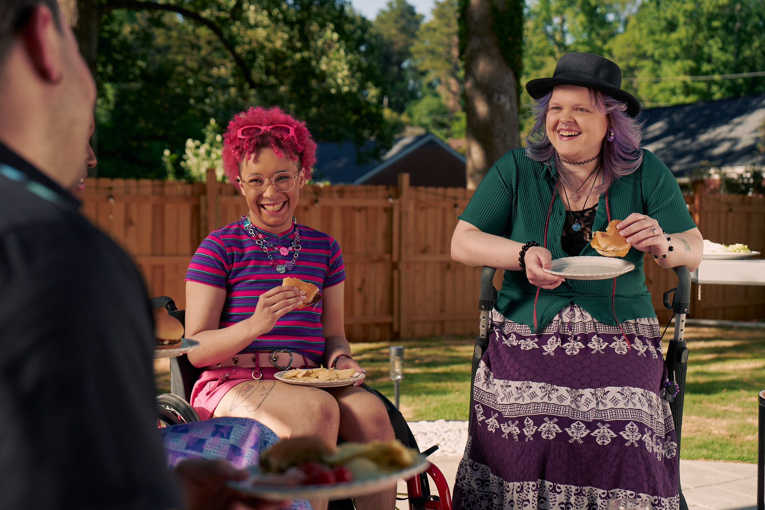 At a backyard barbeque, two nonbinary people sit in their mobility aids, laughing and holding plates of food. One nonbinary person, a Black person with a curly pink fro with loc bangs wearing glasses is seated in a manual wheelchair. They are wearing bright pink sunglassese on their head, a purple and pink striped crop top, pink shorts, and purple sandals. They have several tattoos on both legs. The second nonbinary person, a white person with layered purple hair with two long thin red braids on each side, is seated on a dark gray rollator. They are wearing a black hat, a green shirt, and a purple patterned skirt.