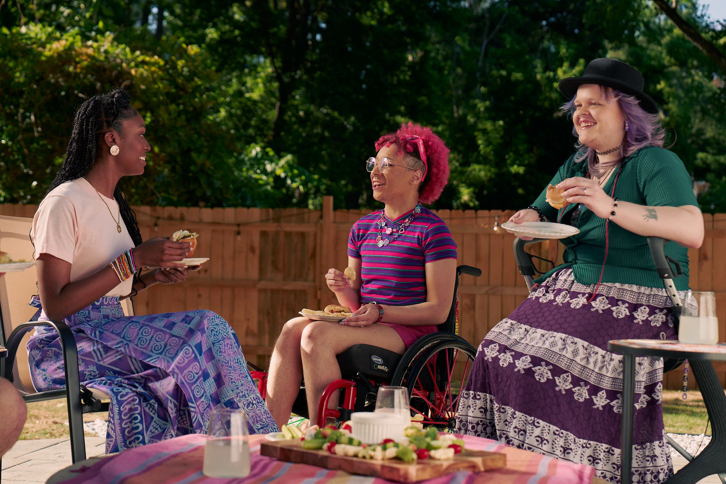 At a backyard barbeque, two nonbinary people sit in their mobility aids, chat with a friend while holding plates of food.  One nonbinary person, a Black person with a curly pink fro with loc bangs wearing glasses is seated in a manual wheelchair. They are wearing bright pink sunglassese on their head, a purple and pink striped crop top, pink shorts, and purple sandals. They have several tattoos on both legs. The second nonbinary person, a white person with layered purple hair with two long thin red braids on each side, is seated on a dark gray rollator. They are wearing a black hat, a green shirt, and a purple patterned skirt. Their friend, an African American woman with long braided hair, is wearing a pink shirt and patterned blue and purple skirt.