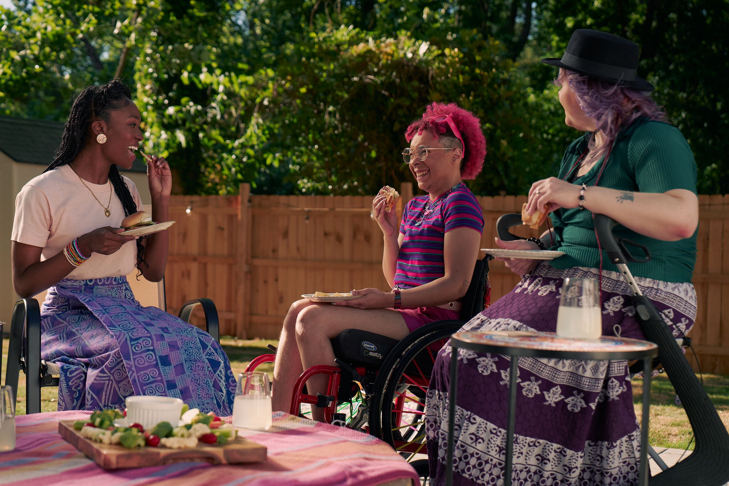 A Black nonbinary person wearing glasses smiles widely while holding a burger. They are seated in a manual wheelchair in a cirle of friends. They have a curly pink fro with loc bangs and are wearing bright pink sunglassese on their head, a purple and pink striped crop top, pink shorts, and purple sandals. They have several tattoos on both legs. Next to them, an African American woman with long braided hair holds a plate of food and smiles. A white nonbinary person seated on a dark gray rollator looks at them. They have purple layered hair with two thin red braids on both sides and are wearing a black hat, green shirt, and purple patterned skirt.