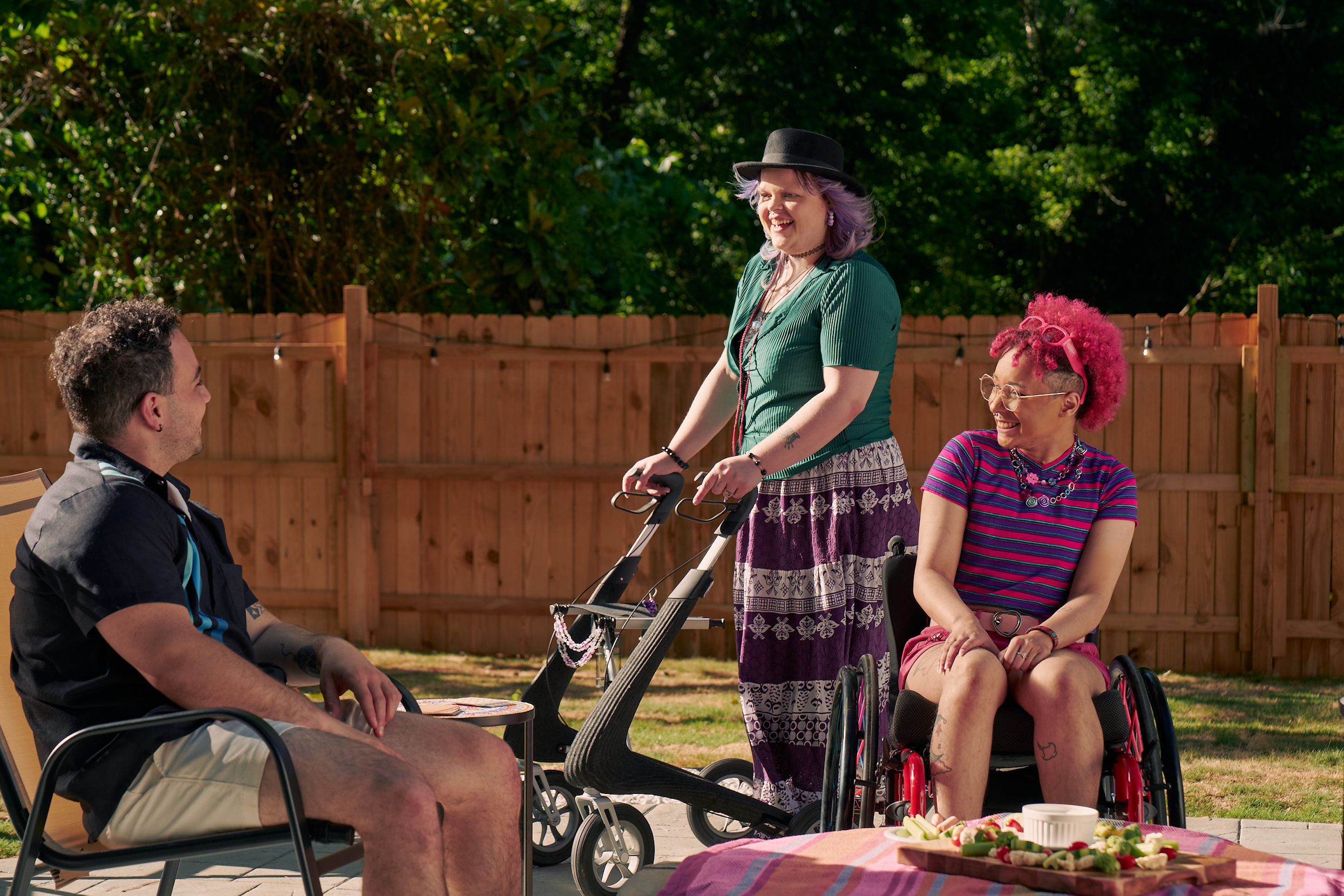 A white nonbinary person stands with their uniquely shaped dark gray rollator that is decorated with purple acrylic chains. They have purple layered hair with two long thin red braids on both sides and are wearing a black hat, green shirt, and purple patterned skirt. They chat with a friend, who is a multi-ethnic man with curly black hair, who is sitting in a patio chair. Next to them is a Black nonbinary person seated in their manual wheelchair with rainbow spokes. They have a curly pink fro with loc bangs and are wearing bright pink sunglassese on their head, a purple and pink striped crop top, pink shorts, and purple sandals. They have several tattoos on both legs.