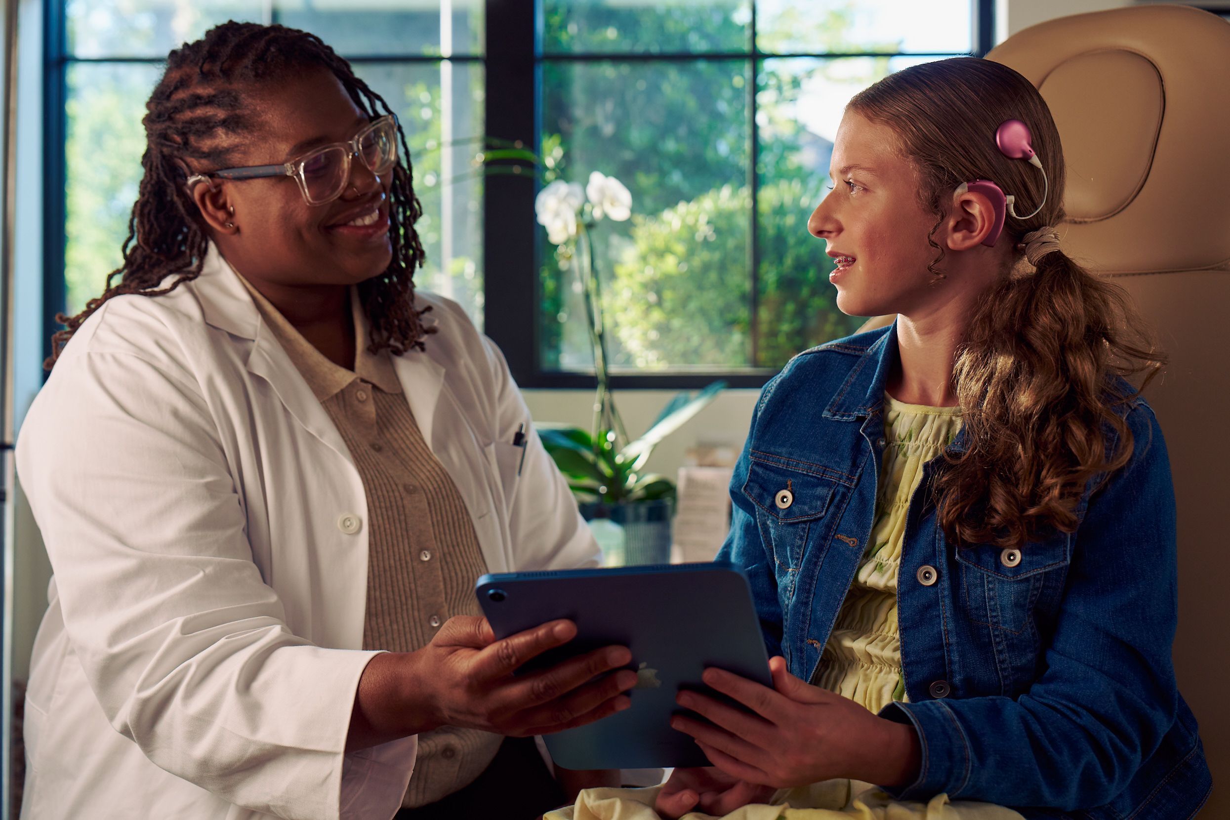 A Black deafBlind audiologist with a cochlear implant and glasses and a young white deaf teen with braces hold a tablet together in a doctor's office. The teen is seated in a medical treatment chair and looks at the audiologist with curiosity. She and has long, wavy sandy blonde hair and is wearing a jean jacket, and a yellow blouse and skirt.