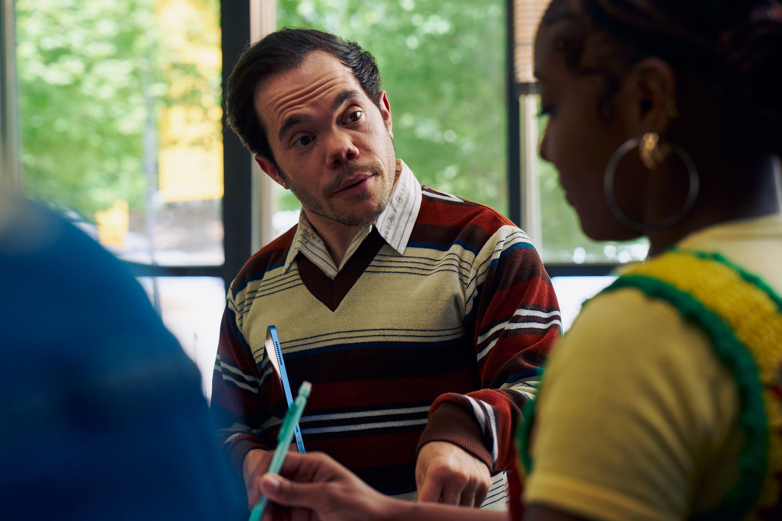 A white male professor with medium brown hair and light brown eyes who has dwarfism stands and talks to a student with focus. He is holding a tablet with one hand and taps the student's desk with the other. The student takes notes as the professor speaks. The professor is wearing a striped V-neck sweater, charcoal dress pants, and dressy-casual shoes. The student is a Black woman with hair pulled into a bun and is wearing a yellow shirt. Behind them are large windows that show a college campus.