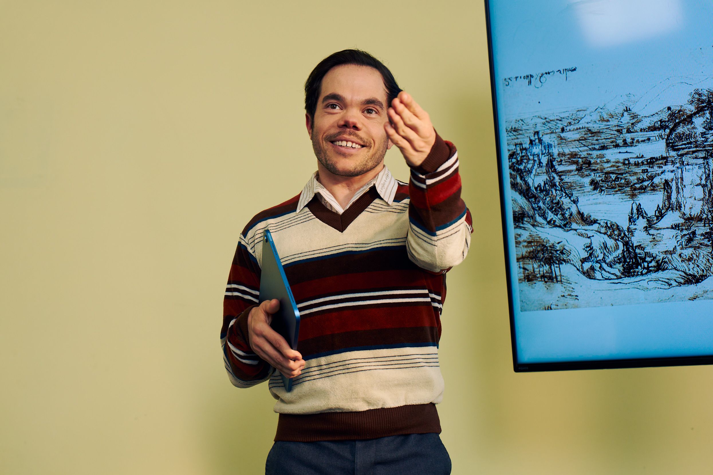 A white male professor with medium brown hair and light brown eyes who has dwarfism smiles and gestures to a student off camera, calling on them. Next to him is a large monitor displaying a pen and ink drawing by Leonardo da Vinci. He is holding a tablet in his other hand. He has short brown hair and is wearing a red striped sweater, a collared shirt, and dark dress pants.