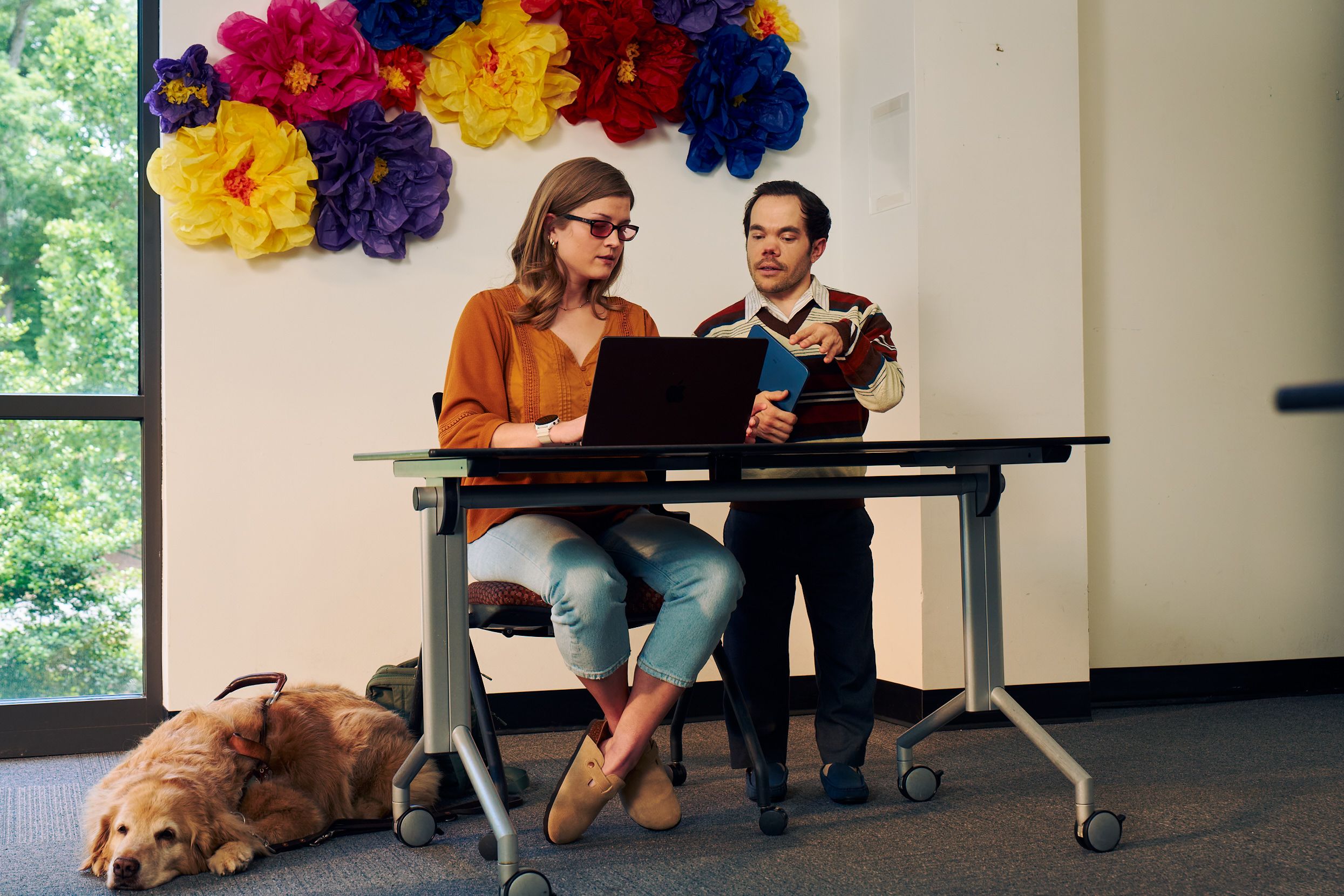 A blind, white woman wearing glasses with red lenses sitting at a lecture table, chats with her professor, a white man with medium brown hair and light brown eyes who has dwarfism, who is standing next to her. They are both looking intently at the student's laptop. A golden retriever service dog wearing a guide harness lays on the floor next to her. The student has dark blonde hair and is wearing an orange blouse and jean capris. A large flower sculpture hangs on the wall behind the lecture table. Next to the sculpture is a large window showing many large trees.