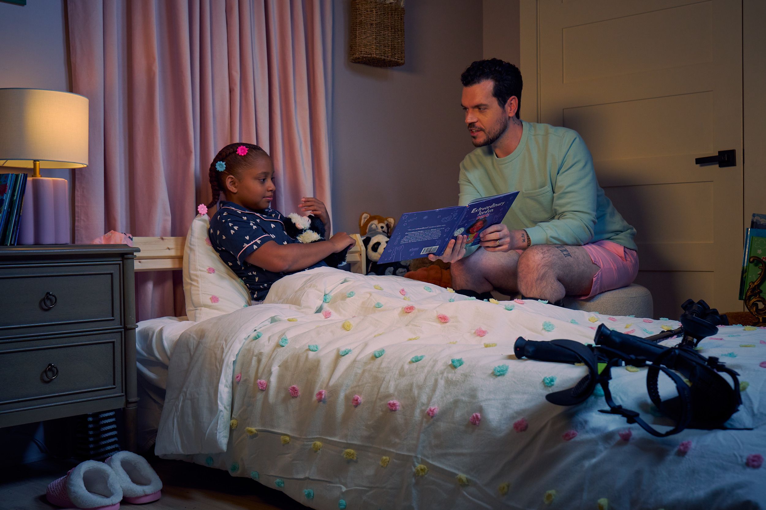 A white man with cerebral palsy who wears leg braces holds a children's book titled "Extraordinary Jordyn and Her Bionic Ears" to his daughter in her bedroom at night. His daughter is a Black multiracial American girl with thick braided red hair adorned with two flower barrettes and is wearing blue patterned pajamas. She is tucked into bed, holding a plushie animal and looks at the open book. The father's black forearm crutches lie on the foot of the bed. He has dark brown curly hair, short brown facial hair, and is wearing a light green sweater with pink shorts. He has a tattoo of the Roman numeral two followed by a slash and the number three. The child's room is painted light pink and includes a nightstand, lamp, a pair of slippers, curtains, and children's books. Her bedspread is white with blue, pink, and yellow polka dots. Framed illustrations of Black women hang on the wall.