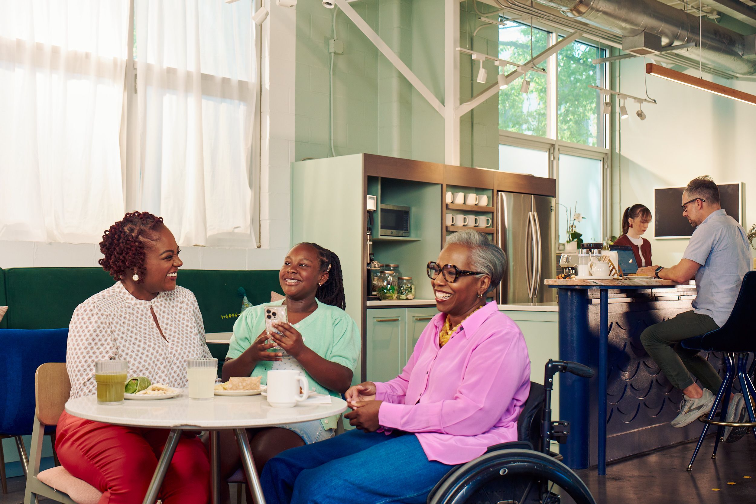 An African American girl shows smiles and holds her phone at a table in a modern cafe, while the girl's mother and auntie laugh together. The auntie is paraplegic and is seated in a manual wheelchair. She has short gray hair is wearing a lavender silk blouse and dark blue jeans. The mother has short tightly curled red hair and is wearing a patterned blouse and red pants. The daughter is wearing a cochlear implant and has her braided hair pulled back into a pony tail. She is wearing a light green cardigan and jean shorts. Behind them, a barista makes drinks and a man types on a laptop.