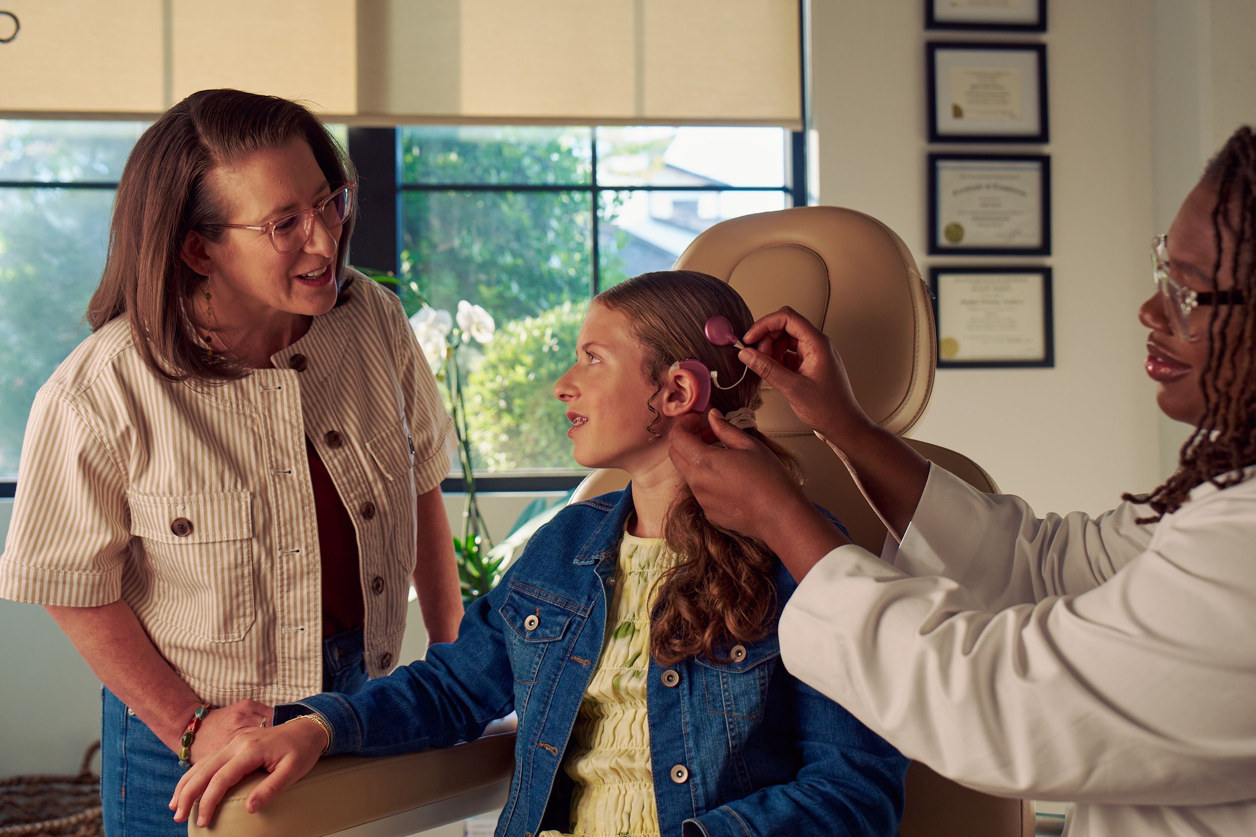 A young white deaf teen with braces sits in a medical treatment chair while an audiologist places a mauve colored cochlear implant on her head. The teen looks at her mother, who is standing and looking at the procedure with curiosity. The teen has long, wavy sandy blonde hair and is wearing a jean jacket, and a yellow blouse and skirt. The audiologist is a Black deafBlind woman wearing glasses and has dark brown shoulder length locs. The teen's mother is a middle-aged white woman with straight brown hair and glasses. She is wearing an off-white striped summer jacket and jeans. A row of four framed professional degrees are hanging on the wall behind them.