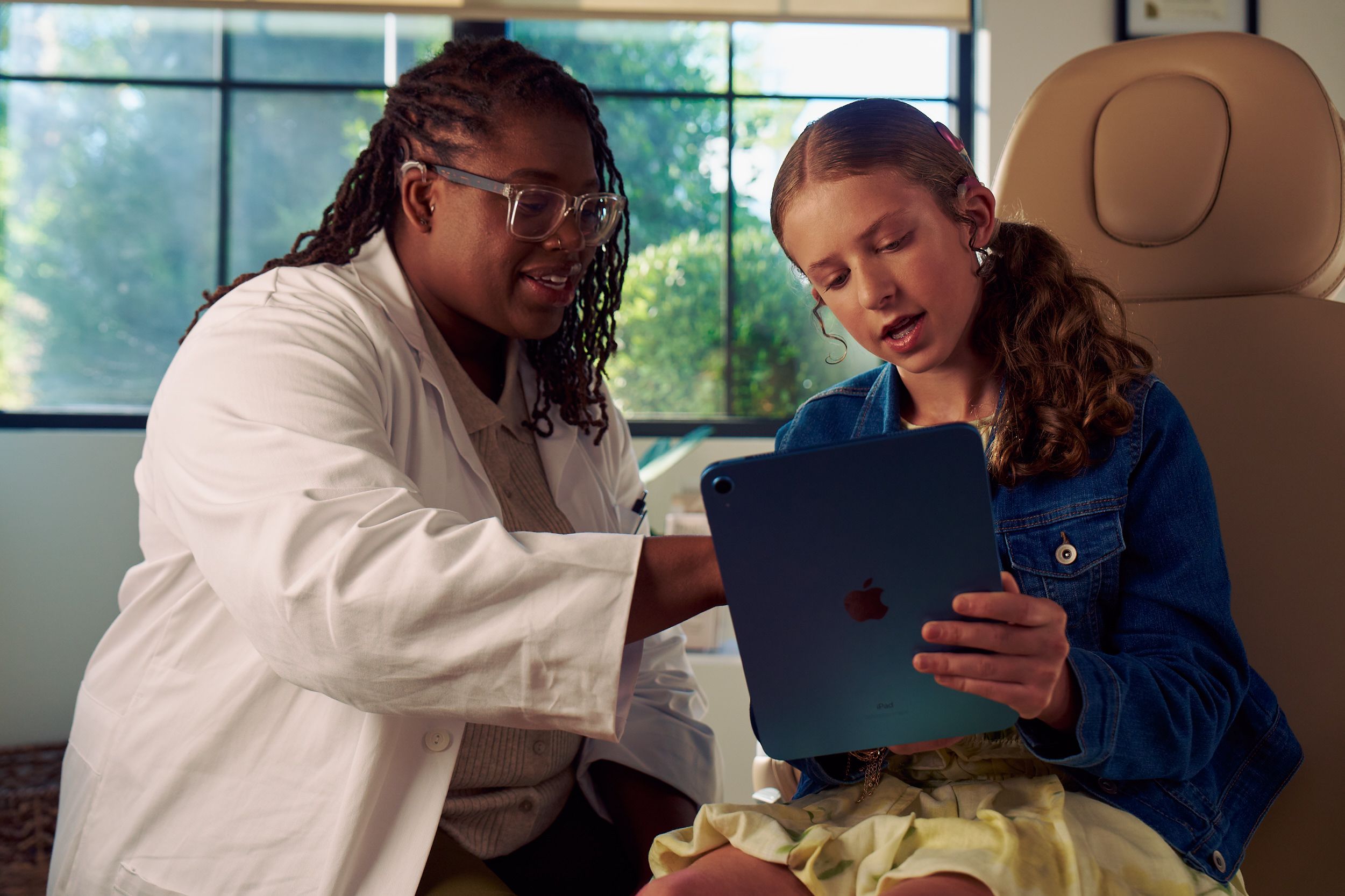 A Black deafBlind audiologist with a cochlear implant and glasses points to something visible on a tablet that a young white deaf teen with braces is holding. The teen is seated in a medical treatment chair and looks at the tablet with curiosity. She and has long, wavy sandy blonde hair and is wearing a jean jacket, and a yellow blouse and skirt.
