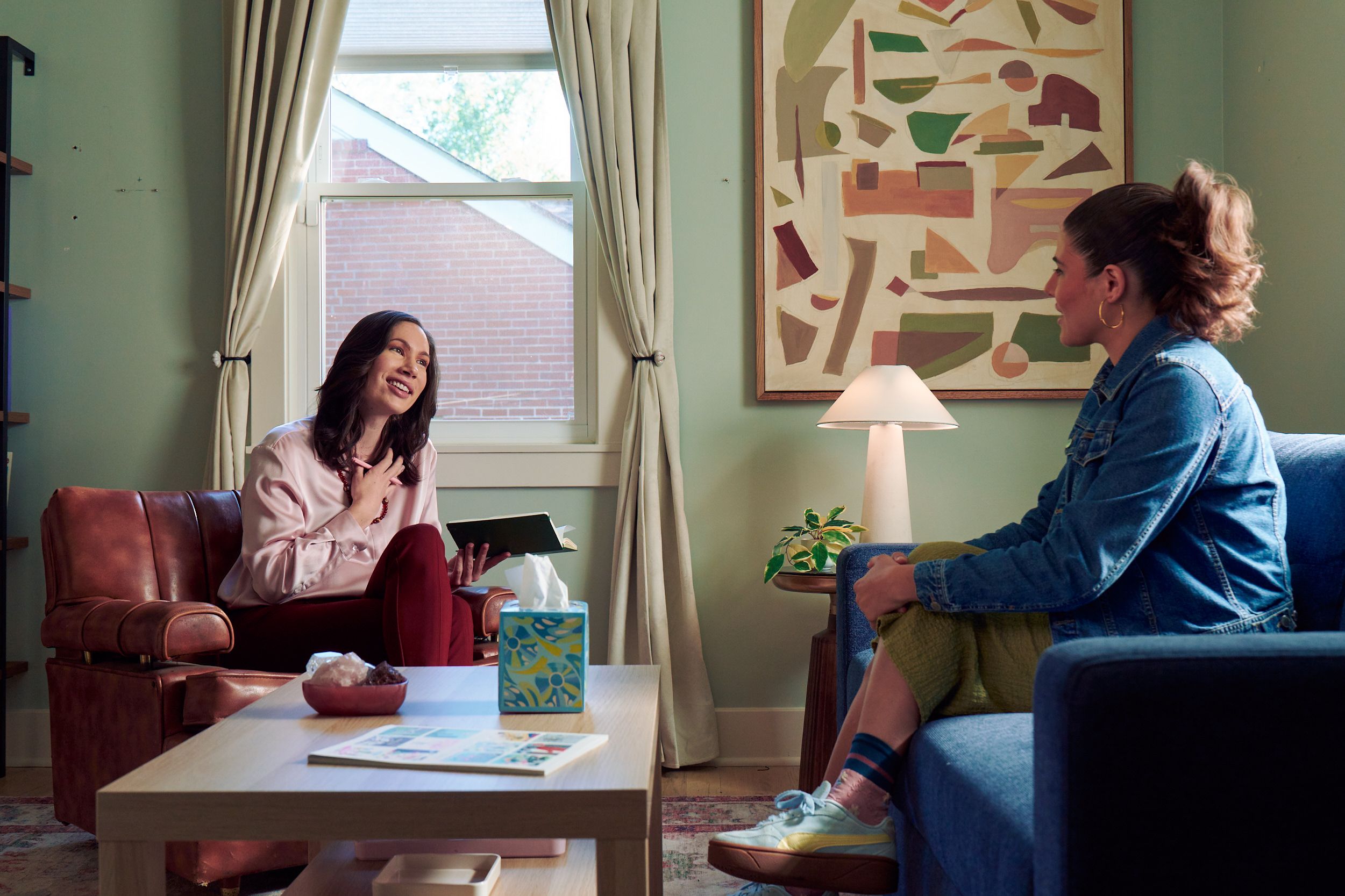 An Eastern European white woman therapist sits in a large chair and looks at a client seated on a couch in front of her. She is holding her hand to her chest and smiles at her client knowingly. The client is a Latina woman with dark brown wavy hair pulled into a ponytail. A tissue box, a red marble heasrt-shaped bowl of crystals, and a magazine are on a low coffee table in the center of the office. An abstract art piece, a lamp, and a small house plant are next and a window letting in natural light. The therapist is wearing a silk pink top, maroon dress pants, and a minimalist necklace. The client is wearing gold hoop earrings, a jean jacket, green skirt, and sneakers.