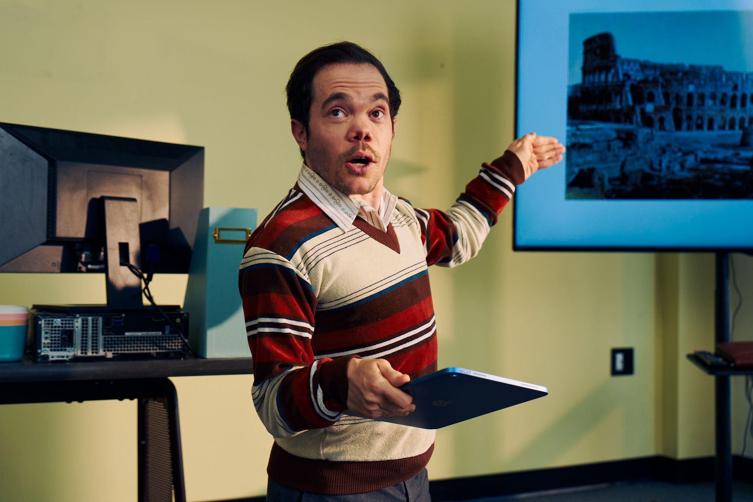 A white male professor with medium brown hair and light brown eyes who has dwarfism gestures at a large monitor next to him, displaying a black and white photo of the Colosseum. He is holding a tablet in his other hand. He is dressed in a striped V-neck sweater, charcoal dress pants, and dressy-casual shoes, standing with a warm, confident presence. Behind him, his desk has a computer, a file holder, and a mug.
