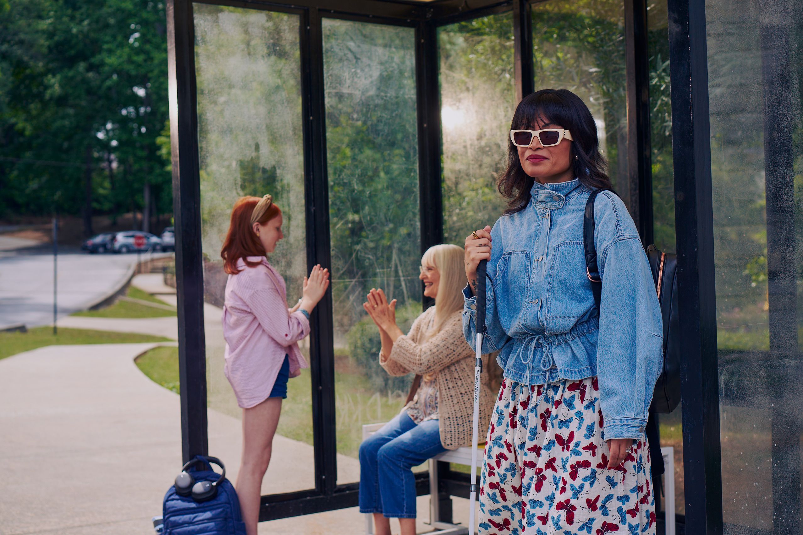 An Asian Filipino blind woman with Retinitis Pigmentosa and Usher Syndrome stands at a covered bus stop. She is wearing white sunglasses and is holding her white cane. She has on a jean jacket, a butterfly patterned skirt, and a black backpack. Behind her, a white European girl with red hair plays pattycake hand game with her grandmother, who is sitting on the bus stop bench. Behind the bus stop are several large trees and walking paths.