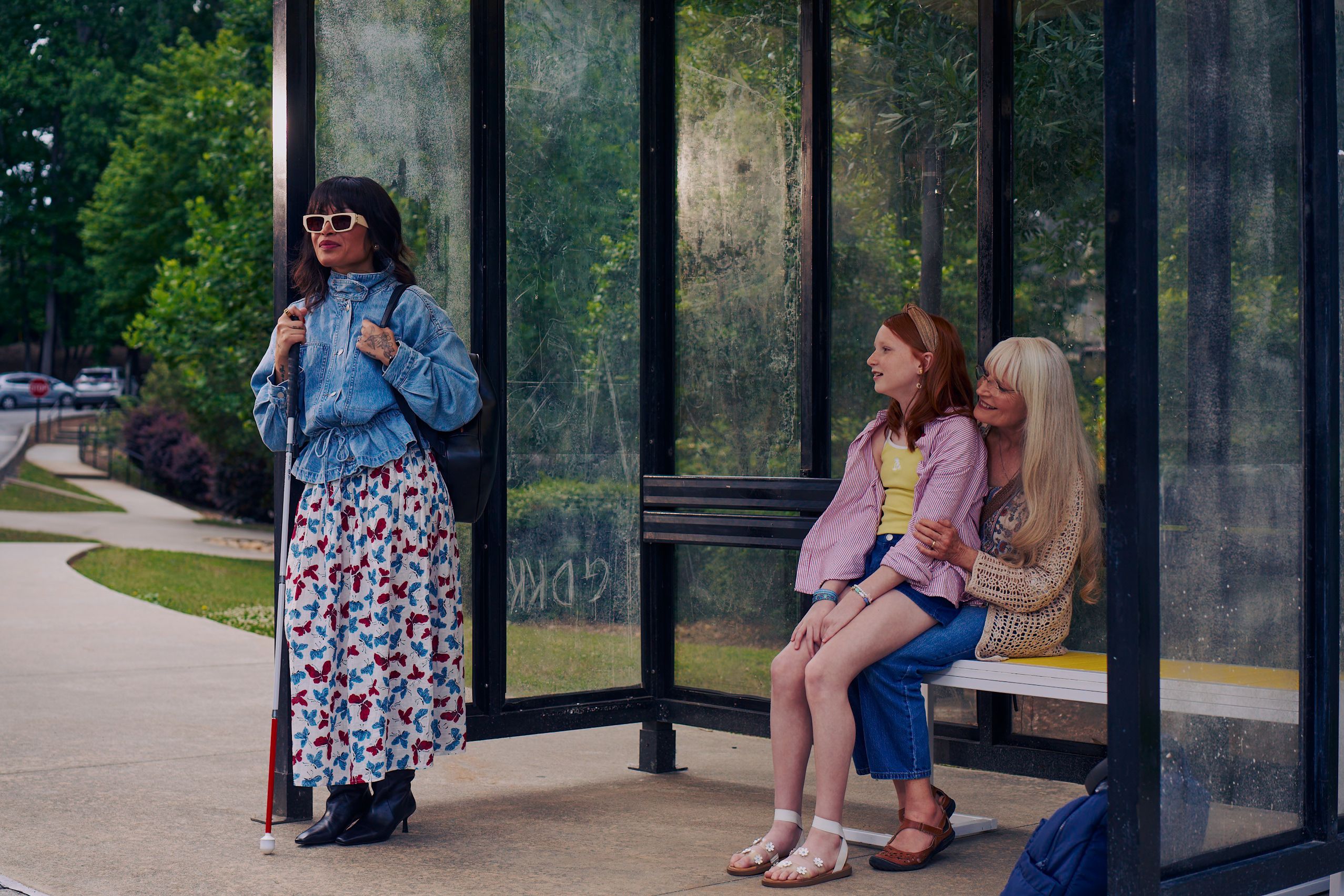 An Asian Filipino blind woman with Retinitis Pigmentosa and Usher Syndrome stands at a covered bus stop. She is wearing white sunglasses and is holding her white cane with her tattooed hands. She has on a jean jacket, a butterfly patterned skirt, and a black backpack. Behind her, a white European girl with red hair sits on her grandmother's lap on the bus stop bench. Behind the bus stop are several large trees and walking paths.