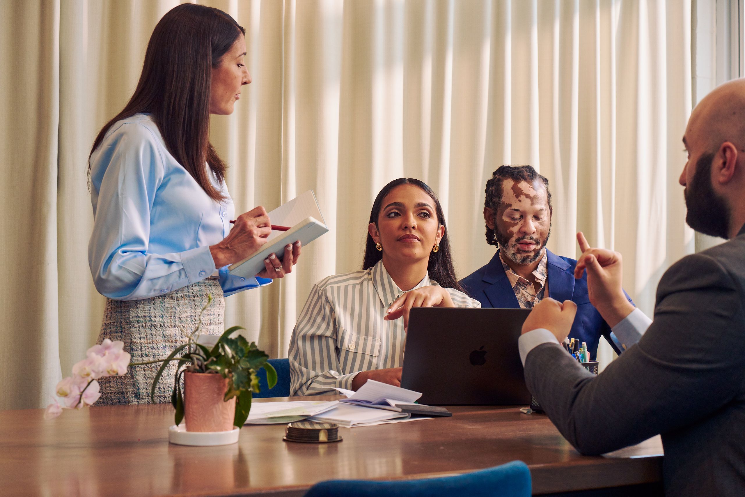 A Caucasian woman with long straight brown hair wearing business attire, holds a notebook and pen while speaking to a Deaf Latina Puerto Rican woman seated next to her. A bald white sign language interpreter sitting across from the women translates the Caucasian woman's speech into American Sign Language, while the Deaf woman looks on. The interpreter is wearing a gray business suit. The Deaf woman has long deep brown hair and is wearing a striped business shirt and hoop earrings. An African American man with olive and porcelain skin, Non-Segmental Vitiligo, and brown barrel-twist dreadlocks wearing a business suit looks downward. A laptop, business planning documents, a cup of pens, and a potted plant are on the conference room table.
