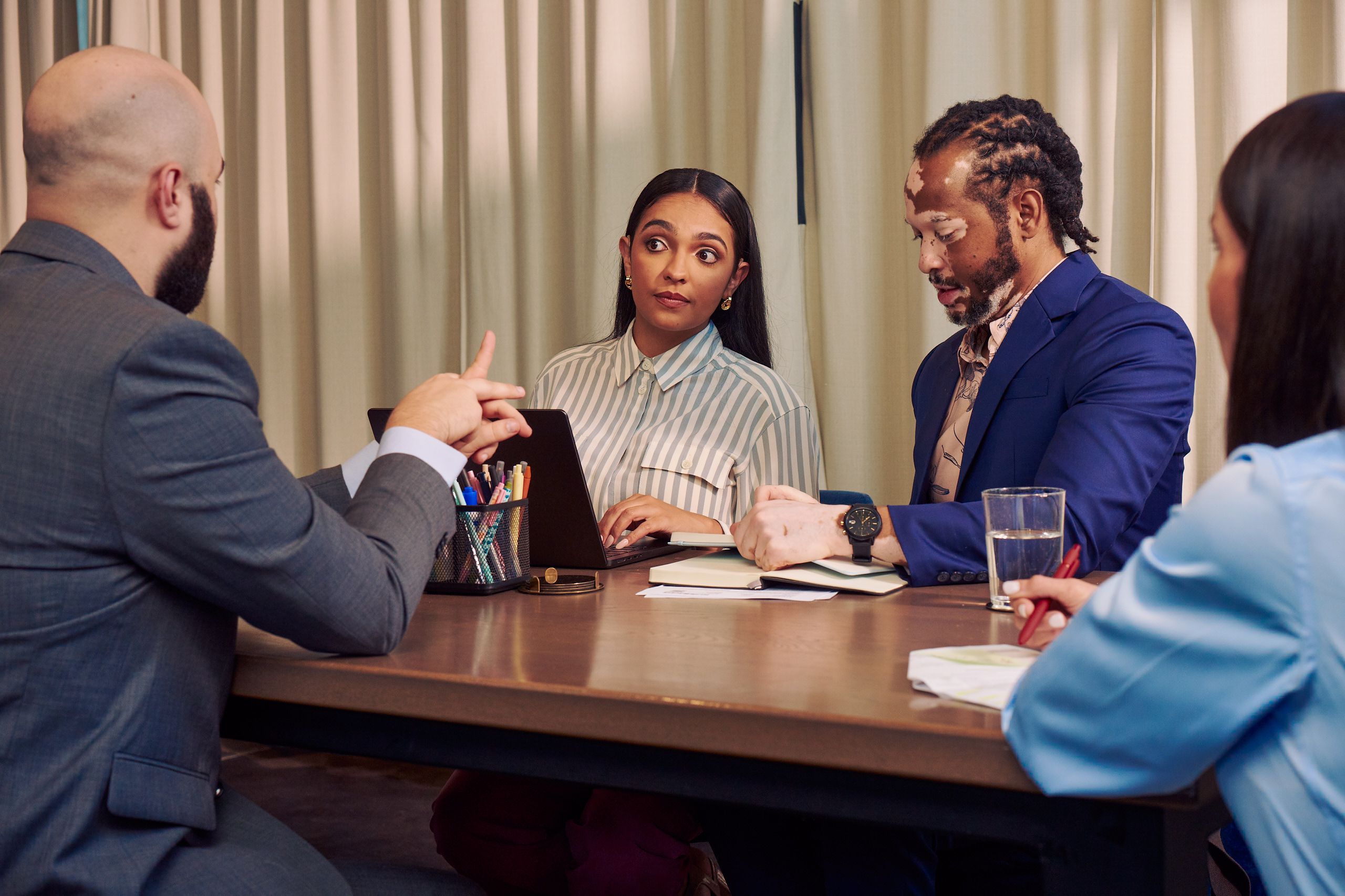 A bald white sign language interpreter translates speech into American Sign Language for a Deaf Latina Puerto Rican woman sitting across from him at a conference room table. An African American man with olive and porcelain skin and Non-Segmental Vitiligo is seated next to her at the table, writing in a notebook. A Caucasian woman with long brown hair writes in a notebook across the table. All four colleagues are wearing business attire.