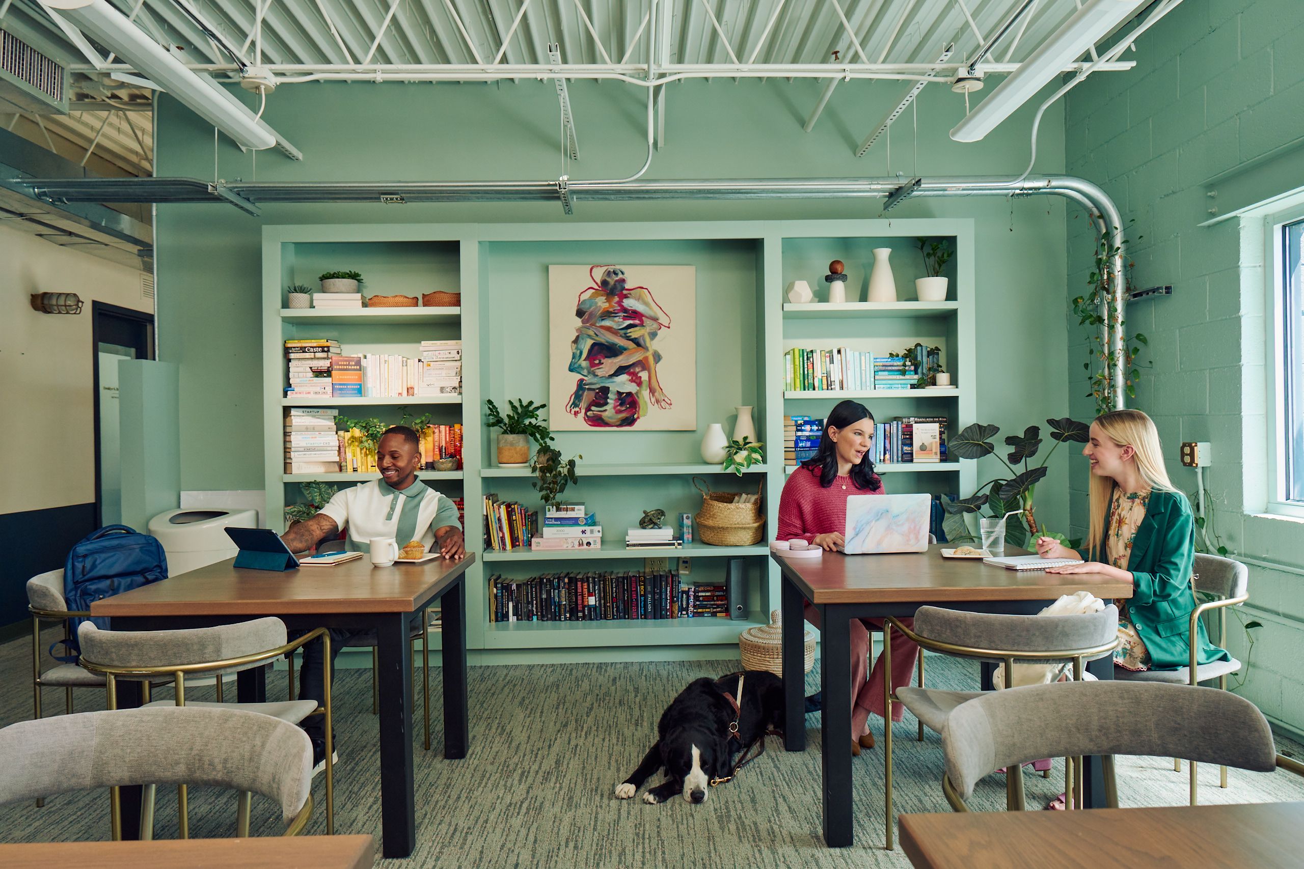 Colleagues chat and work in a modern coworking office space. At one table, a young white woman with partially paralyzed arms and hands chats with a young pale white blind woman. A black and white guide dog with a guide harness lies on the floor next to the blind woman, who is typing on a laptop computer with headphones nearby. She has shiny black hair styled in waves and is wearing a chunky knitted pink sweater and dusky pink flared pants. The white woman with partially paralyzed arms and hands is smiling while writing in a notebook, with a glass of water with a straw in it nearby. She has long, straight blonde hair and is wearing a floral dress and a green blazer. At another table, a Black man smiles while looking at his tablet computer. He is wearing a white and green polo shirt and a pastry and mug are nearby. The coworking office space is painted a warm mint green and has a large bookcase displaying several books, plants, art pieces, and clay vases.