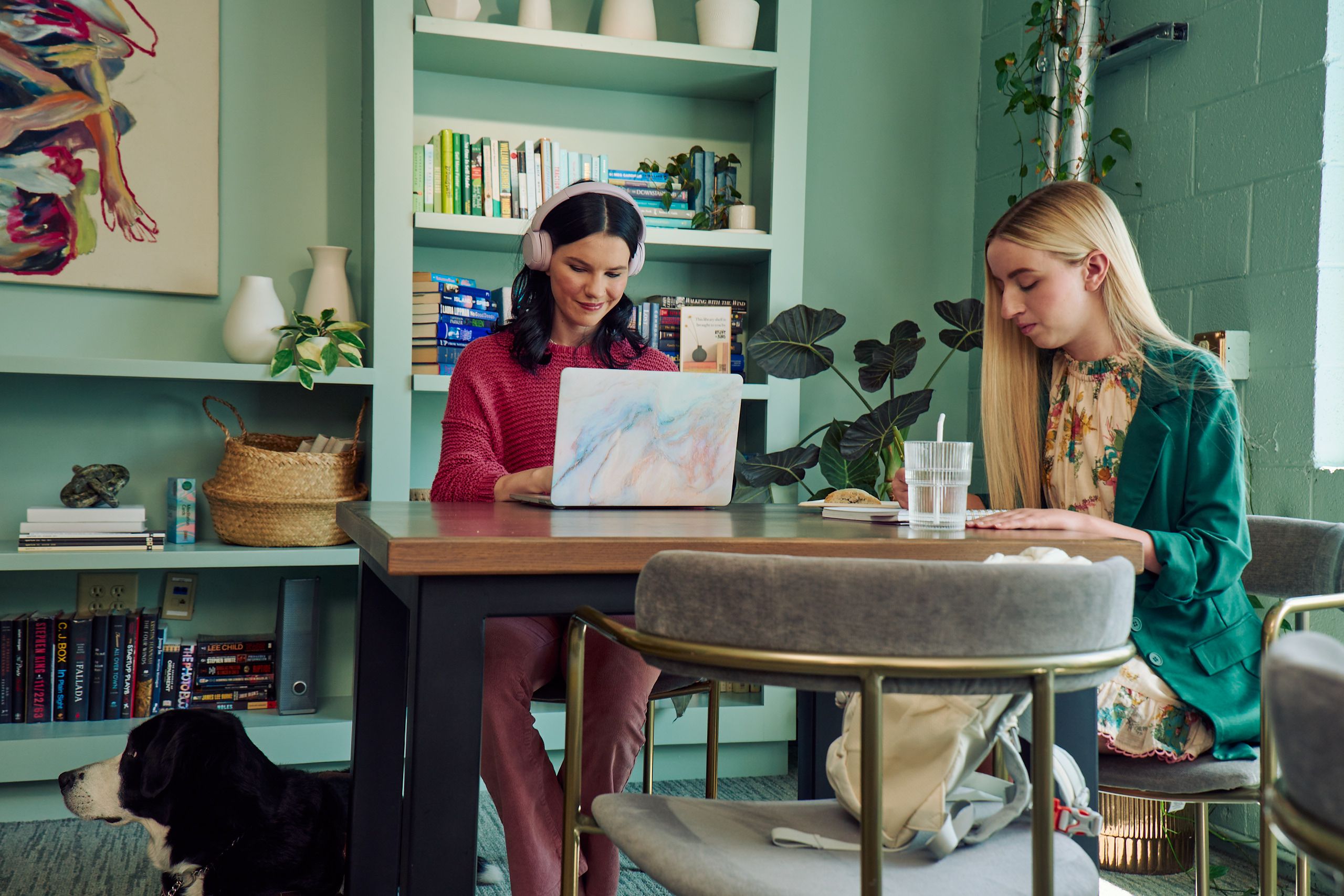 A young pale white blind woman and a young white woman with partially paralyzed arms and hands are coworking at a table in a modern coworking office space. The blind woman types on a computer while the woman with partially paralyzed arms and hands writes in a notebook. A glass of water with a straw in it is nearby. The woman with partially paralyzed arms and hands has long straight blonde hair and is wearing a floral dress and green blazer. The blind woman has shiny black hair styled in waves and is wearing a chunky knitted pink sweater and dusky pink pants. Her black and white guide dog is near her feet. The coworking office space is painted a warm mint green and features a large bookcase with several books, plants, clay vases, and a large abstract art piece.