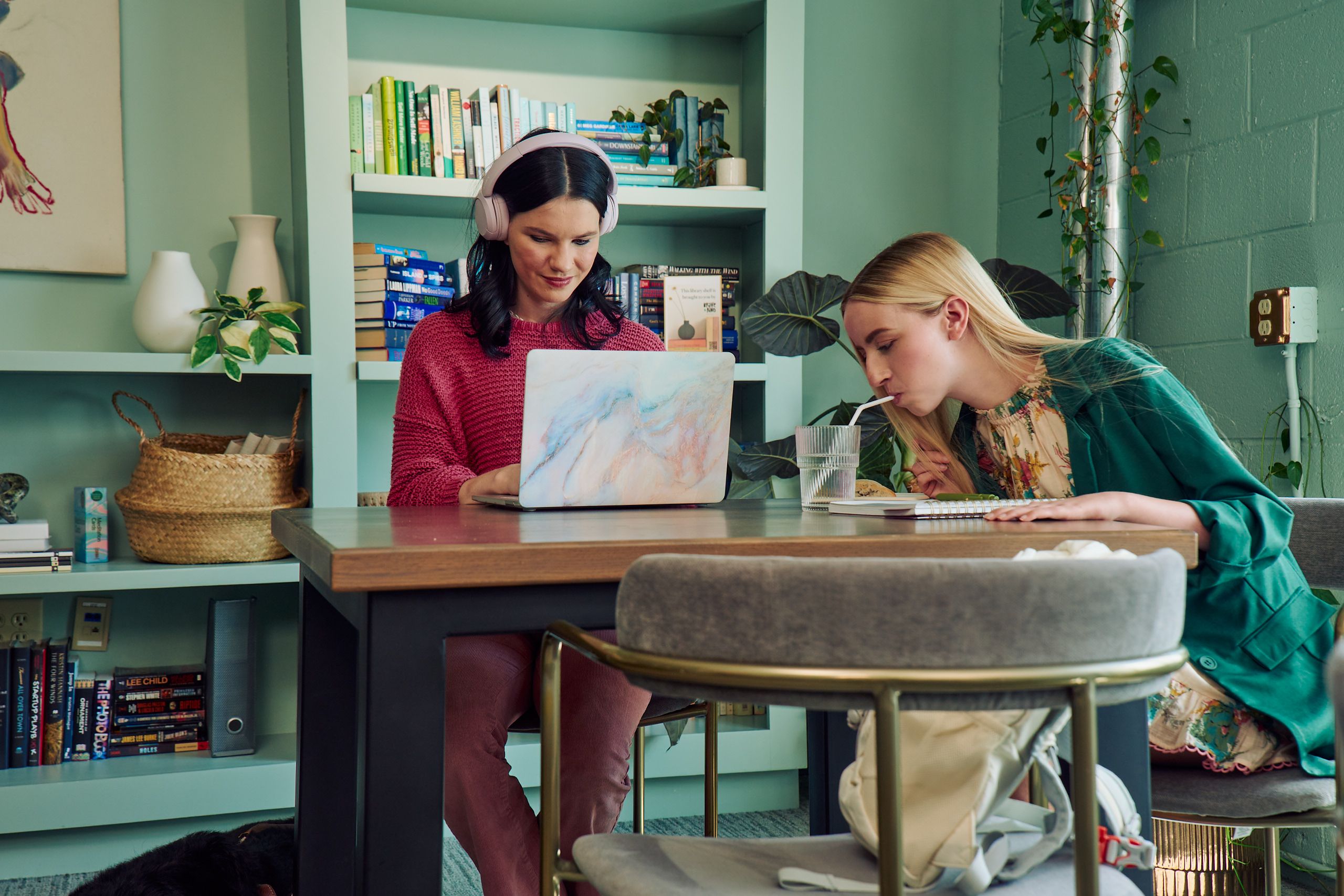 A young white woman with long, straight blonde hair and partially paralyzed arms and hands bends forward to drink water from a cup through a straw. She is sitting at a table in a modern coworking office space with a young pale white blind woman, who is typing on a laptop computer. The blind woman has shiny black hair styled in waves and is wearing over the ear headphones, a chunky knitted pink sweater, and dusky pink flared pants. The woman with partially paralyzed arms and hands is wearing a floral dress with pink trim and a green blazer. The coworking office space is painted a warm mint green and has a large bookcase and features several books, plants, a rattan basket, and clay vases.