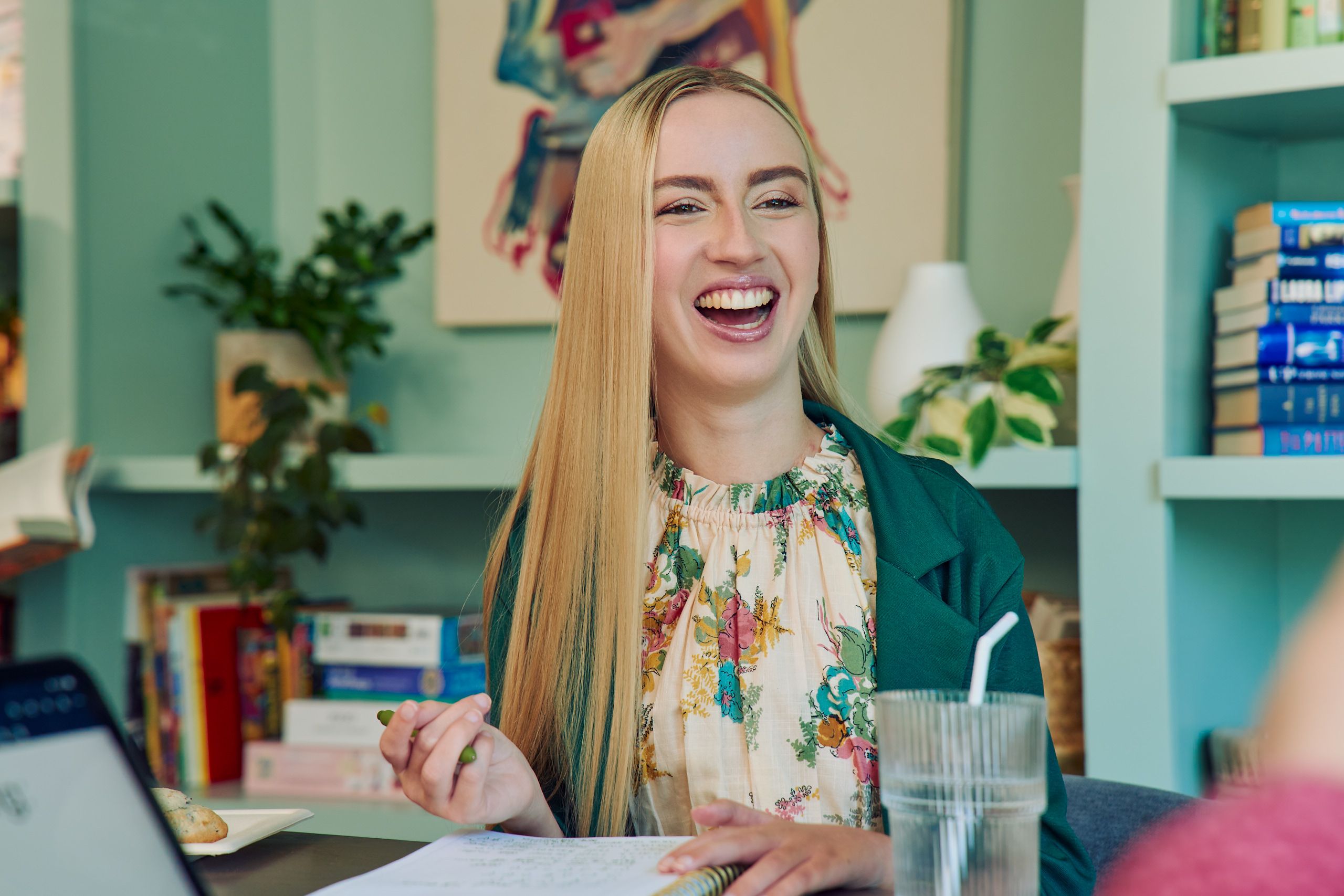 A young white woman with long, straight blonde hair and partially paralyzed arms and hands laughs with a wide smile. She is sitting at a table in a modern coworking office space holding a pen with an open notebook in front of her. A glass of water with a straw in it is nearby. She is wearing a floral dress and a green blazer. The coworking office space is painted a warm mint green and features a large bookcase with several books, plants, and a large abstract art piece.