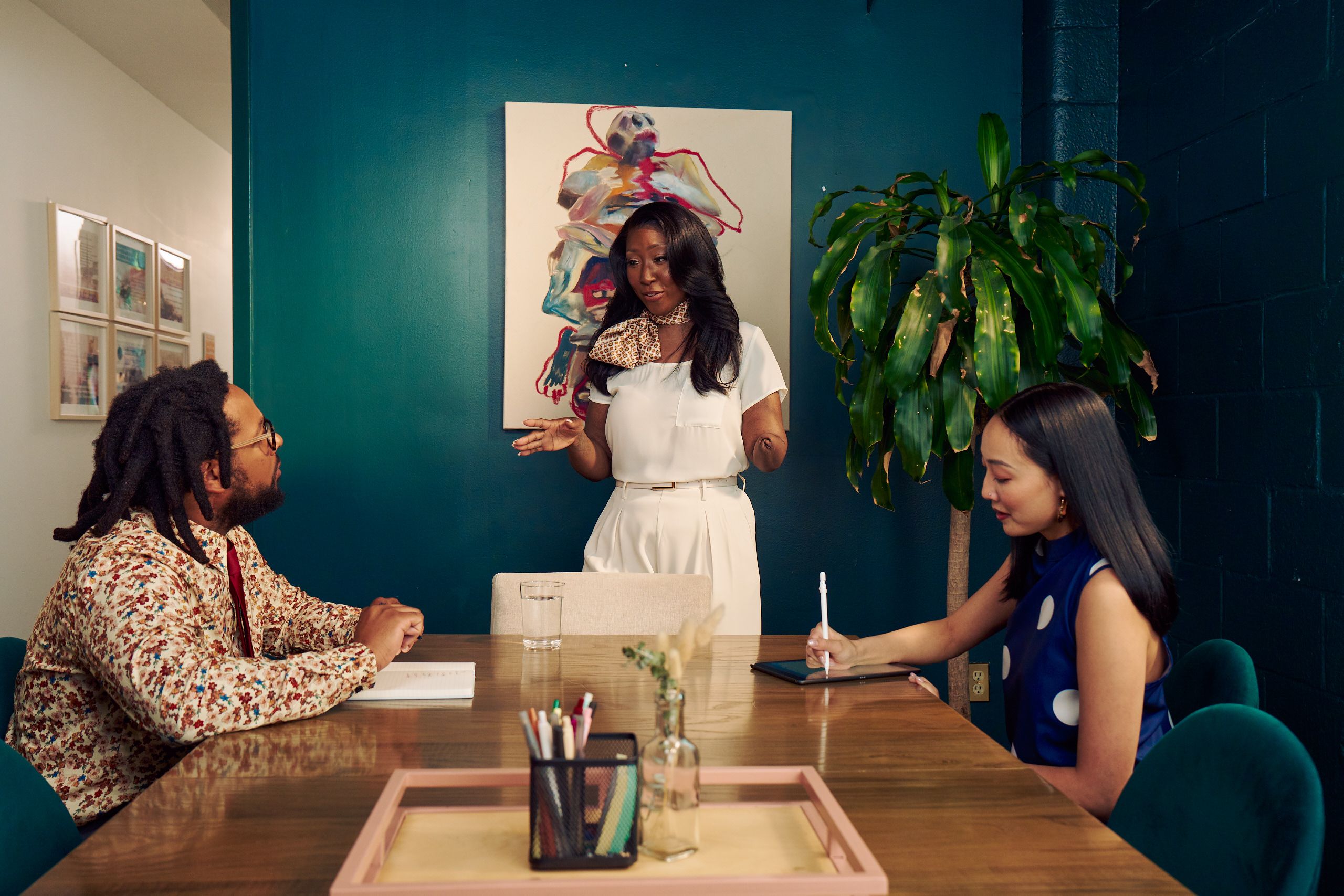 An executive leads a small team meeting in a dark conference room, looking at a neurodivergent Black male sitting at the conference table in front of her. The executive is a Black American woman and below-elbow congenital amputee wearing a cream blouse and pants, with a patterned scarf around her neck. Her hair is side parted and coiffed. The Black male has a notebook in front of him. He has locs and is wearing glasses, a floral print button down shirt, and a red tie. Across the table sits a Korean and Filipino woman wearing a blue polka-dot blouse, smiling. Her tablet is in front of her. The conference room is painted dark teal and features an abstract art piece and a large potted plant. A tray with a cup of pens and a small flower vase is in the center of the conference table. The conference room door is open to a long white hallway with framed art pieces hanging.