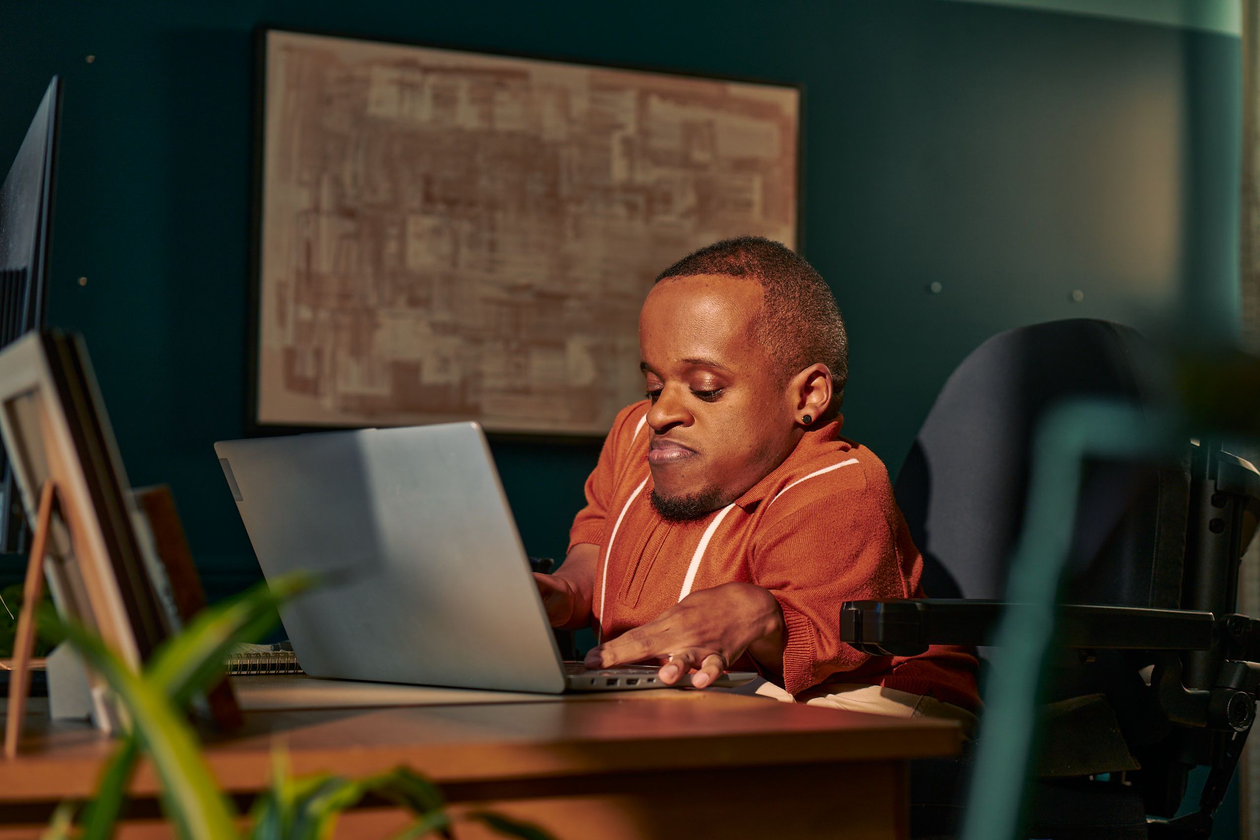 A black man who is a little person is seated in his power wheelchair in a dark teal home office. He types on a laptop on his desk, with a monitor and framed photos nearby. An abstract art piece is on the wall behind him. He has closely cropped black hair, brown eyes, and is wearing a rust-orange knit quarter-zip sweater with white vertical stripes, light khaki trousers, and a small stud earring.