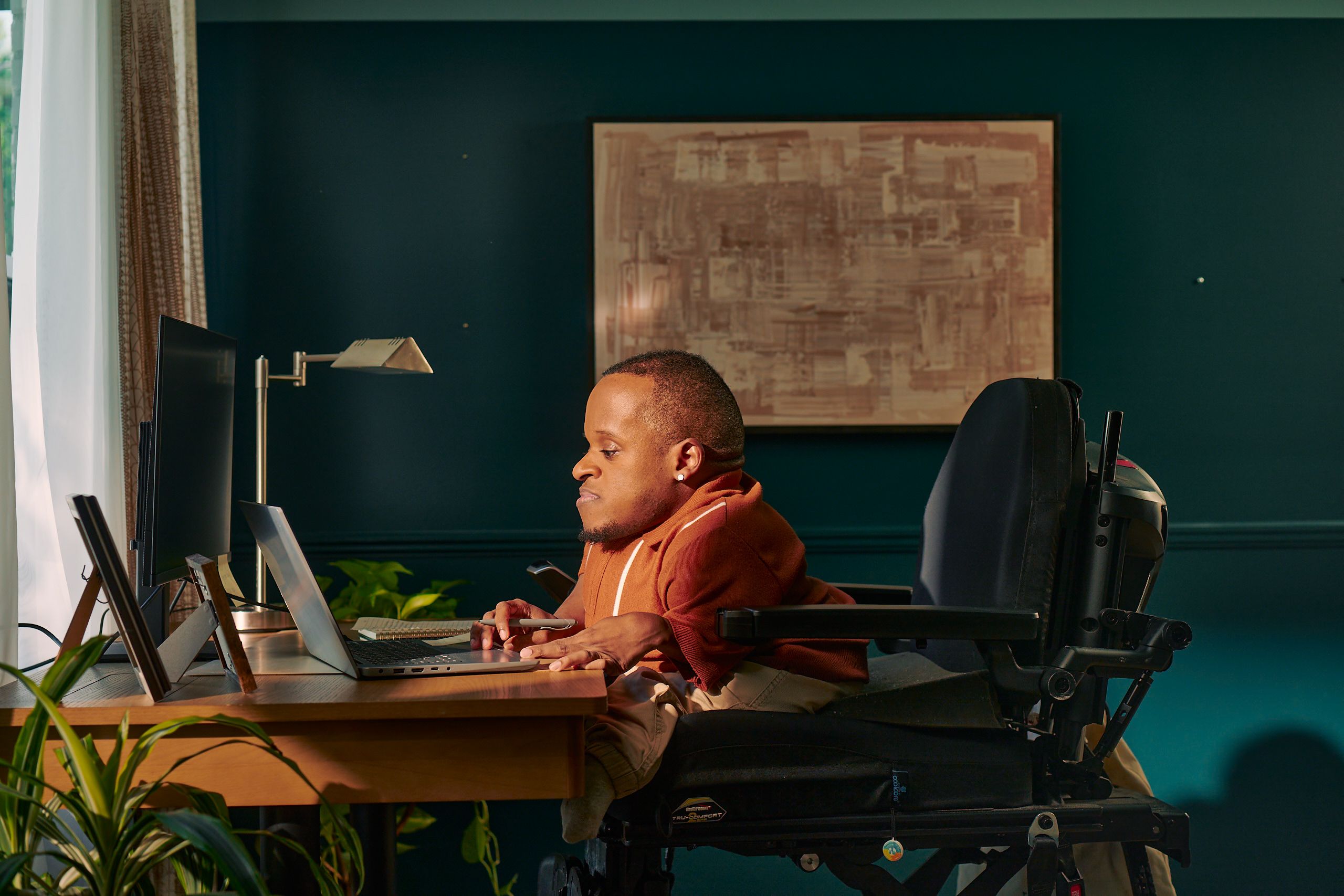 A black man who is a little person is seated in his power wheelchair in a dark teal home office. He holds a pen and types on his laptop. A monitor, notebook, pen, lamp, and framed photos are on the desk in front of him. His wheelchair is in an elevated position, allowing him to be level with his desk. He is facing a window, letting in light through white and patterned curtains. His desk is surrounded by several house plants and an abstract art piece. He has closely cropped black hair, brown eyes, and is wearing a rust-orange knit quarter-zip sweater with white vertical stripes, light khaki trousers, and a small stud earring. A tote bag hangs on the back of his wheelchair.