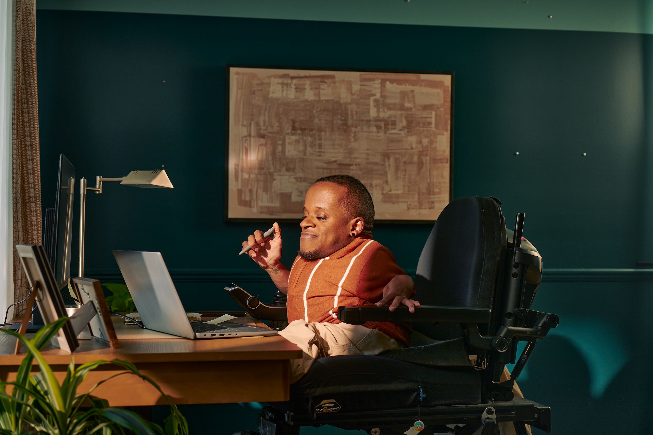 A black man who is a little person is seated in his power wheelchair in a dark teal home office. He holds a pen and looks down at his laptop. A monitor, notebook, pen, lamp, and framed photos are on the desk in front of him. His desk is surrounded by house plants and an abstract art piece. He has closely cropped black hair, brown eyes, and is wearing a rust-orange knit quarter-zip sweater with white vertical stripes, light khaki trousers, and a small stud earring. A tote bag hangs on the back of his wheelchair.