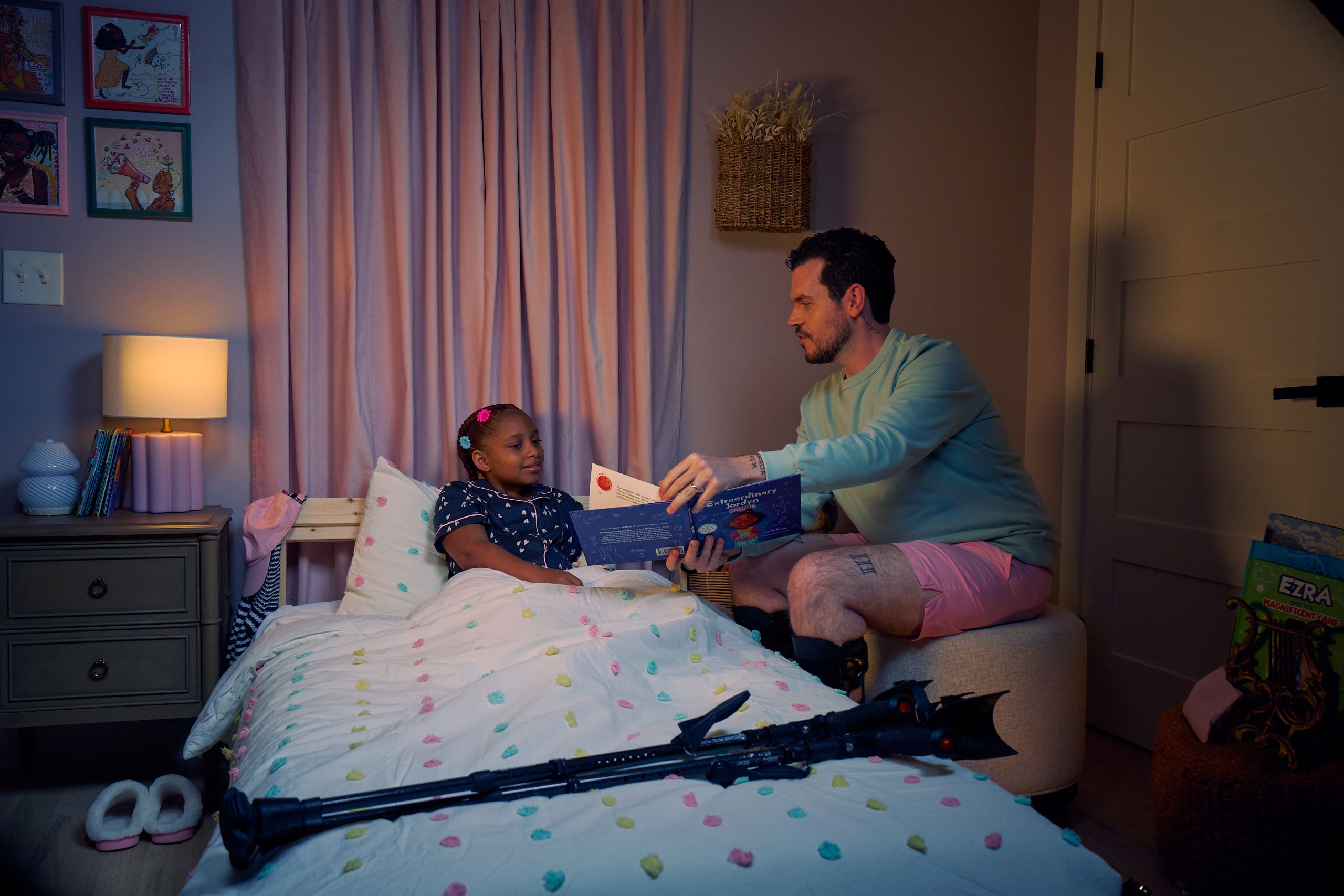 A white man with cerebral palsy who wears leg braces shows a children's book titled "Extraordinary Jordyn and Her Bionic Ears" to his daughter in her bedroom at night. His daughter is a Black multiracial American girl with thick braided red hair adorned with two flower barrettes. She is tucked into bed and is wearing dark blue patterned pajamas and looks at the open book. The father's black forearm crutches lie on the foot of the bed. He has dark brown curly hair, short brown facial hair, and is wearing a light green sweater with pink shorts. He has a tattoo of the Roman numeral two followed by a slash and the number three. The child's room is painted light pink and includes a nightstand, lamp, a pair of slippers, curtains, and children's books. Her bedspread is white with blue, pink, and yellow polka dots. Framed illustrations of Black women hang on the wall.