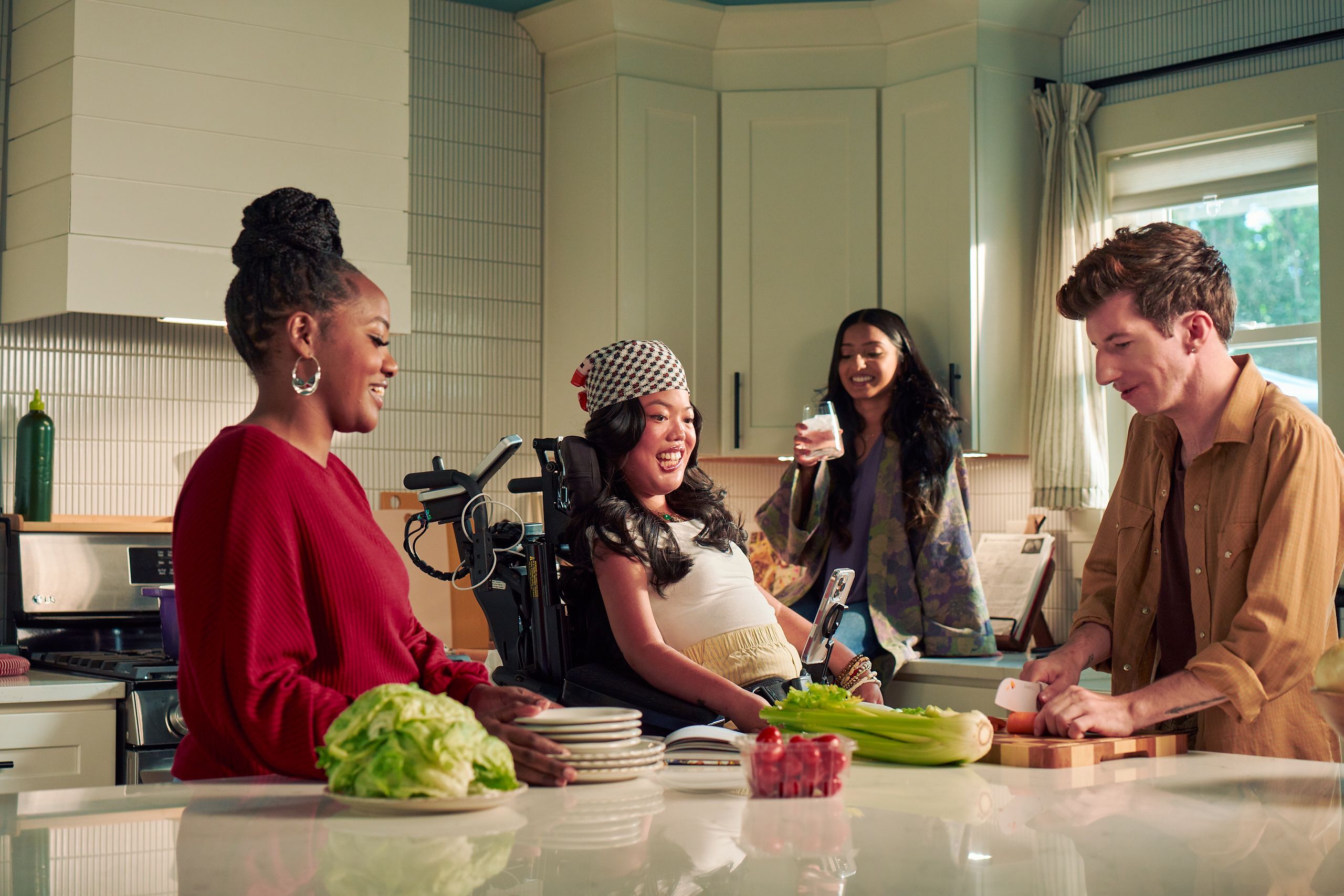 A Southeast Asian woman with curled black hair seated in her power wheelchair smiles and watches her roommate chop vegetables in their bright kitchen. Her smart phone is mounted to her wheelchair. Her roommate is a white male with Moebius Syndrome and a face that is paralyzed. The Southeast Asian woman is wearing a white top with striped, yellow linen pants and a checkered headscarf. He has short, wavy, reddish brown hair and is wearing a tan button down shirt with the sleeves rolled up, showing arm tattoos. Two other roommates laugh and hang out nearby. One roommate has long dark hair and is wearing a purple floral dress, the other is a black female with a braided bun and maroon top.