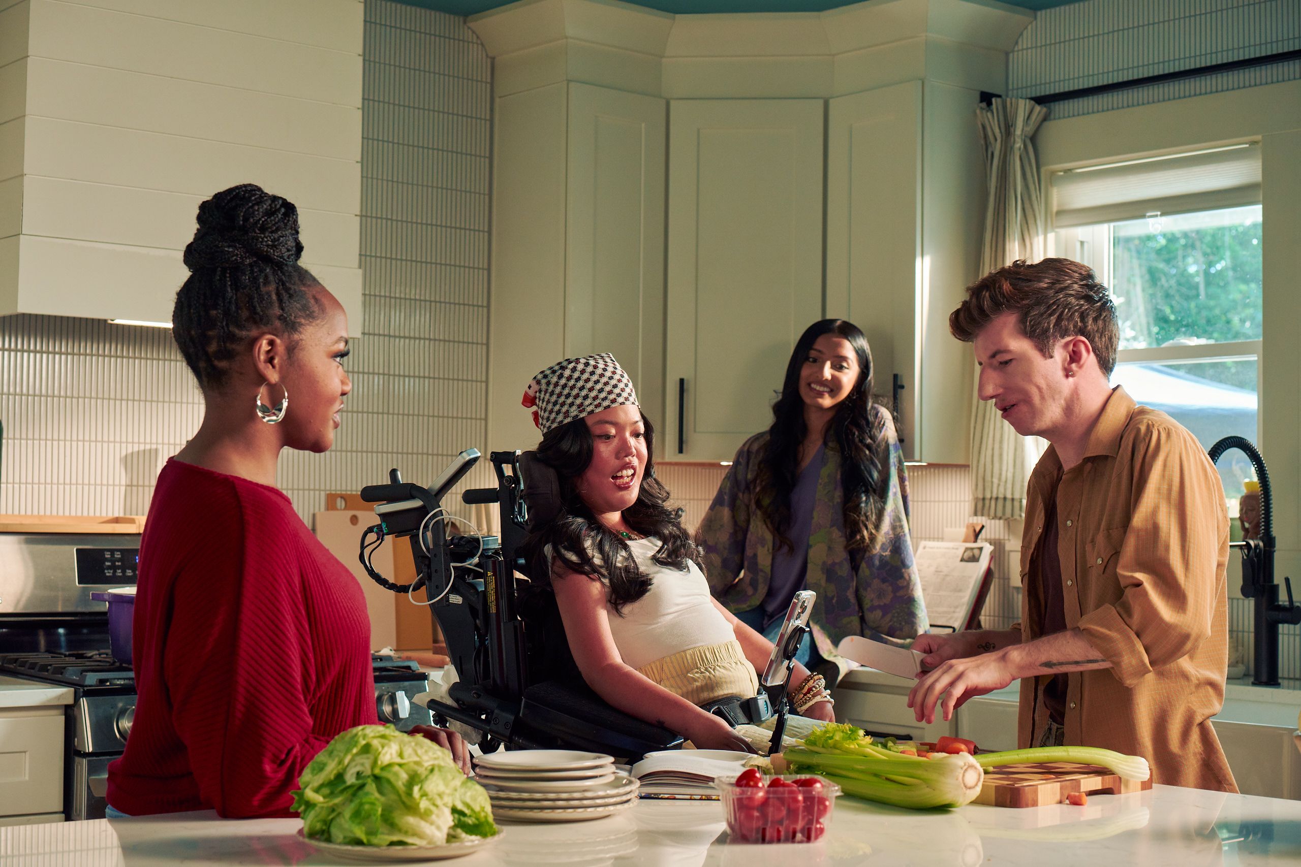 Four roommates hang out in their bright kitchen. One roommate, a white male with Moebius Syndrome and a face that is paralyzed, chops vegetables. Another roomate, a Southeast Asian woman with curled black hair seated in her power wheelchair, watches intently. A third roommate with dark hair wearing a purple floral dress sits on a counter and smiles while watching him prepare the vegetables. A fourth roommate, a black female with a braided bun wearing a maroon top, chats with him.
