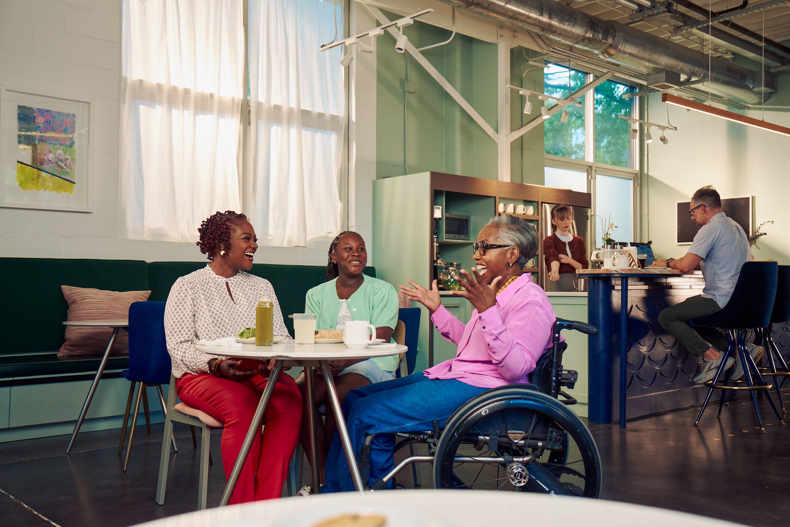 An African American mother, daughter, and auntie are smiling and chatting at a table in a modern cafe. The auntie is paraplegic and is seated in a manual wheelchair. She is smiling wide and raises her arms in excitement. She has short gray hair and is wearing a lavender silk blouse and dark blue jeans. The mother has short tightly curled red hair and is wearing a patterned blouse and red pants. The daughter is wearing a cochlear implant and has her braided hair pulled back into a pony tail. She is wearing a light green cardigan and jean shorts. Behind them, a barista prepares drinks and a man types on a laptop.