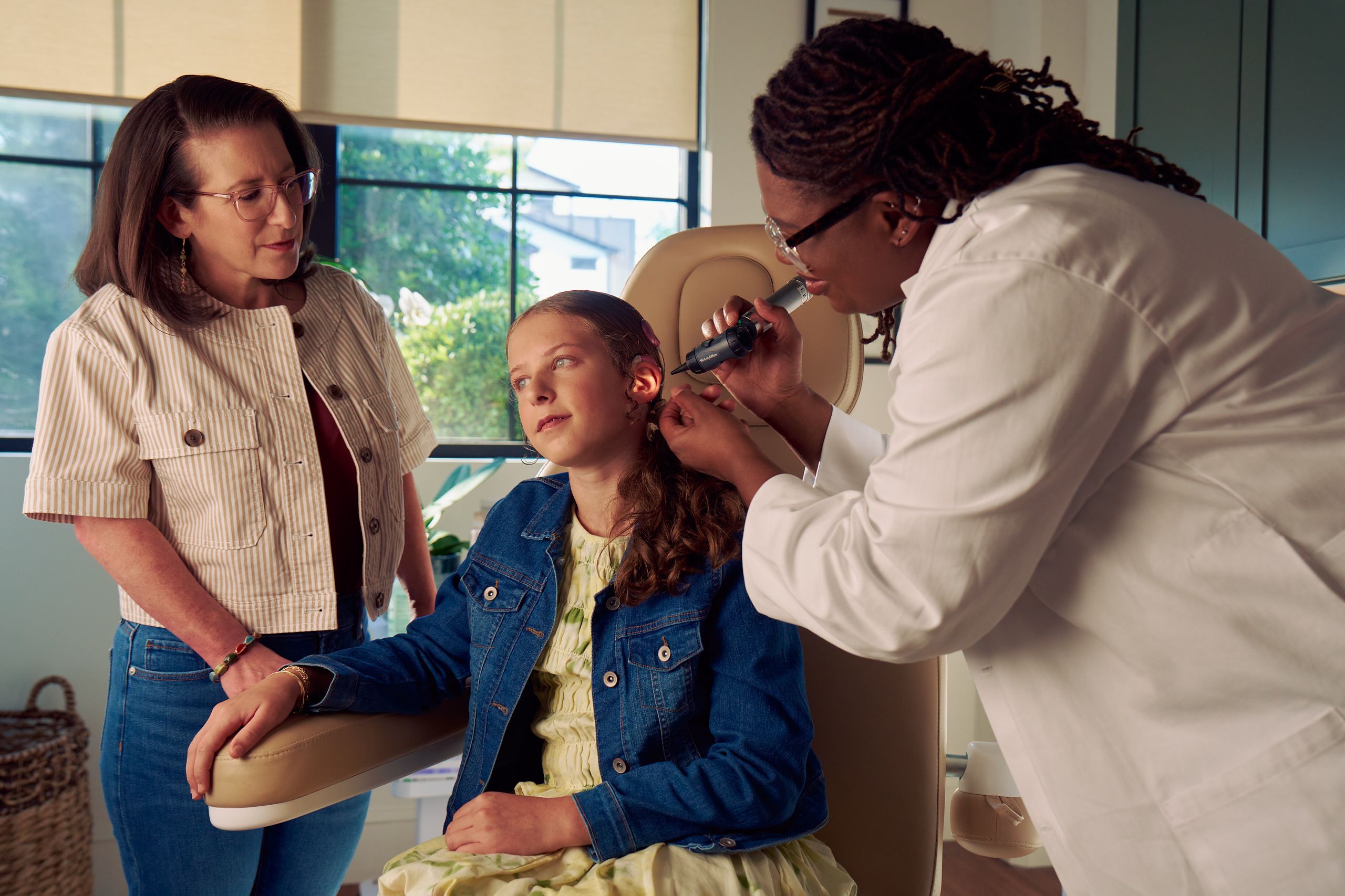 A Black deafBlind audiologist with a cochlear implant and glasses smiles and uses an otoscope to look into the ears of a young white deaf teen with braces seated in a medical treatment chair. The teen is softly smiling and has long, wavy sandy blonde hair and is wearing a jean jacket, and a yellow blouse. The teen's mother is standing next to her, observing the audiologist use the tool.