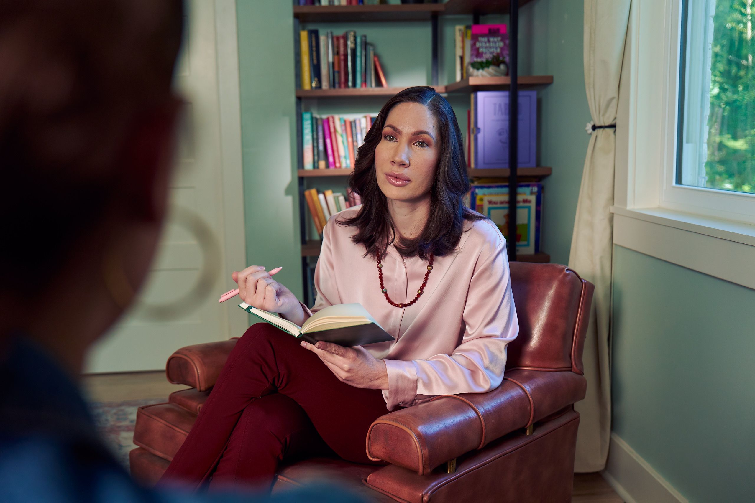 An Eastern European white woman therapist sits in a large chair with an understanding look on her face. She is holding a pen and open notebook, and is looking at a client off camera. The therapist's office has several books on display and a window letting in natural light. The therapist is wearing a silk pink top, maroon dress pants, and a minimalist necklace.