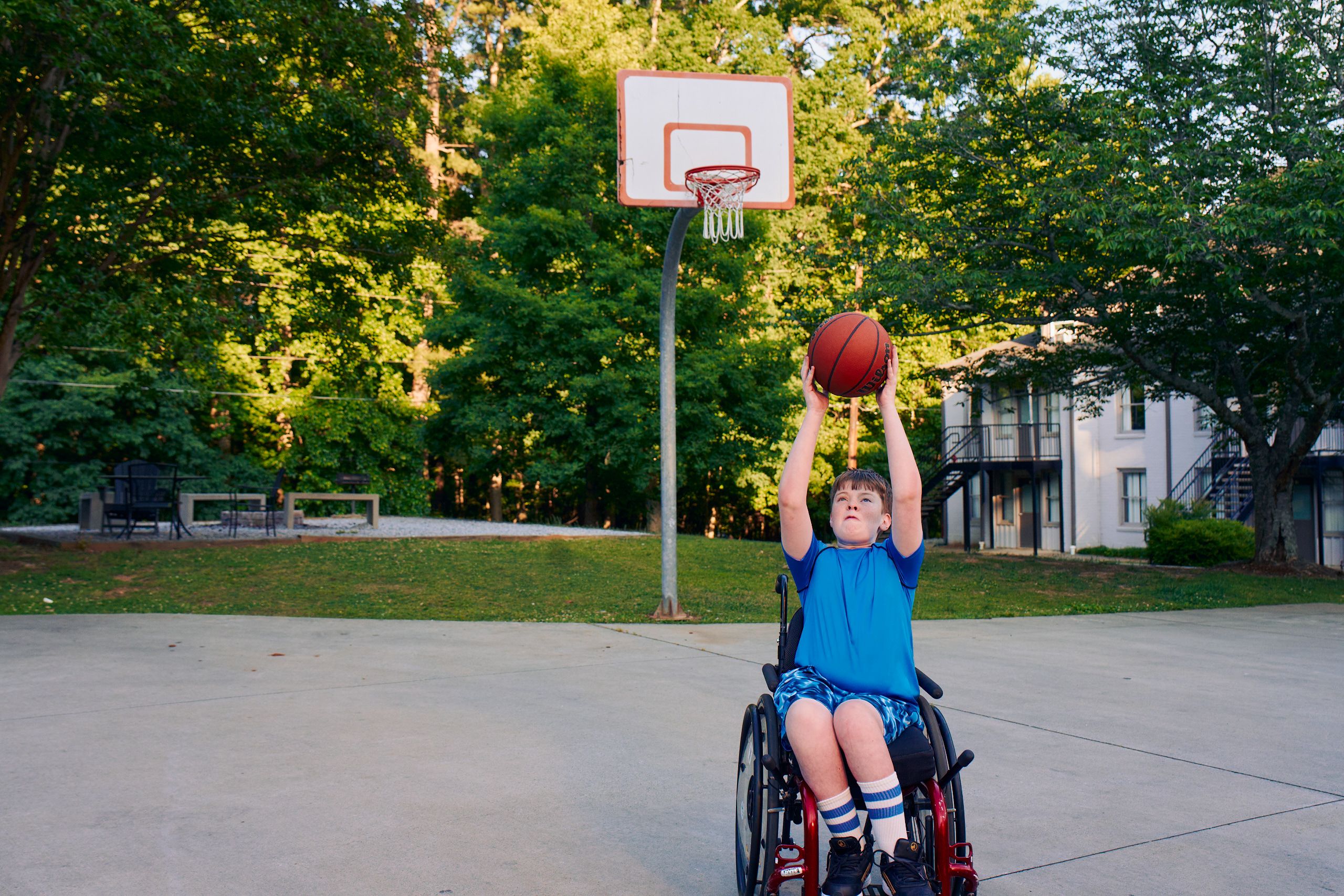 A white teen boy seated in a manual wheelchair holds a basketball over his head, with a look of concentration on his face. He is playing at a basketball court at a neighborhood park. He has short brown hair and is wearing blue athletic wear.
