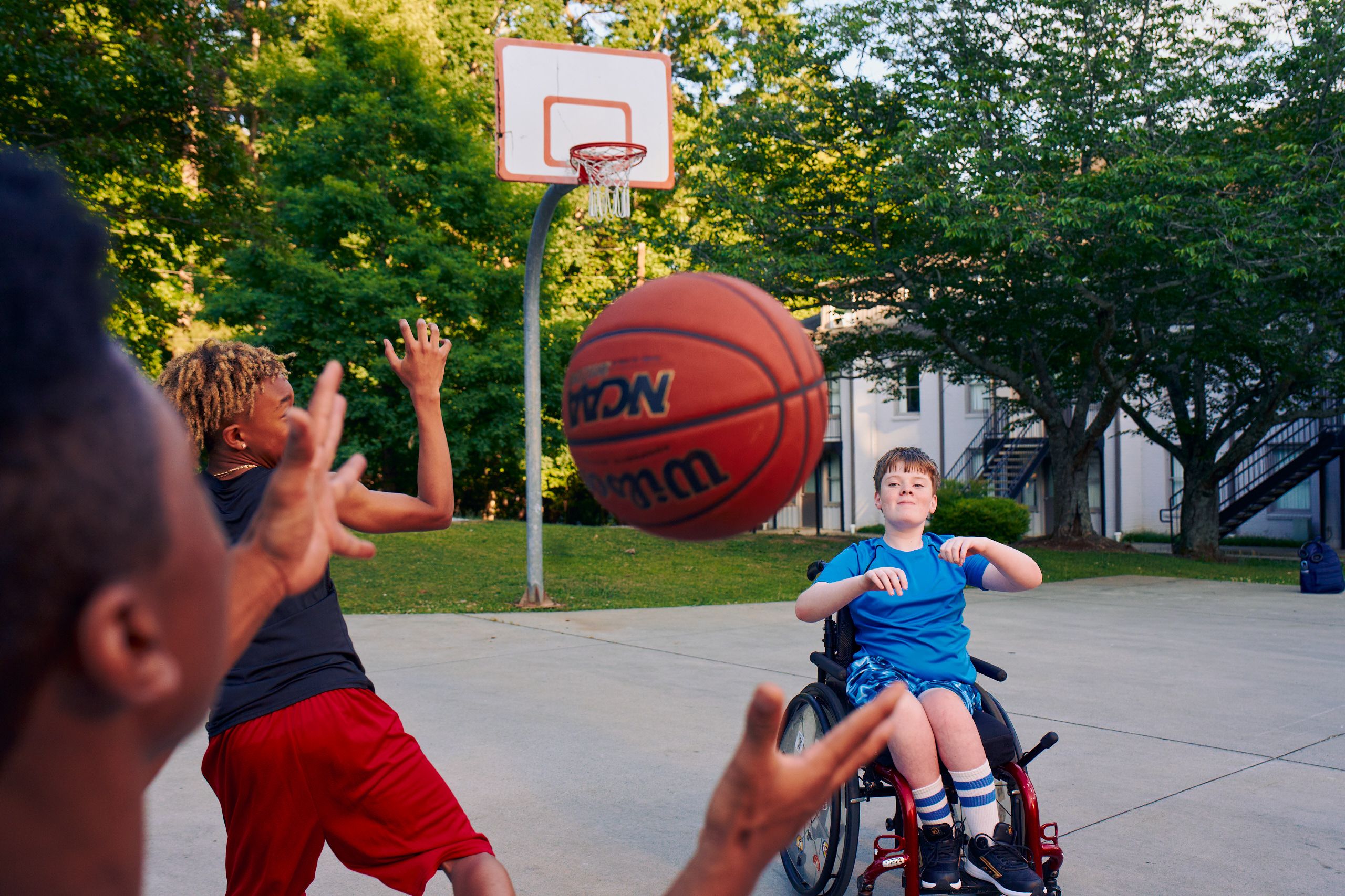 Three teenage friends play basketball at a neighborhood park. One of the teens, a Black boy with short natural black hair styled high on top, passes the ball to another teen, a white boy seated in a manual wheelchair. A third teen, a light-skinned African American male, runs towards the hoop. The teen seated in the wheelchair has a gentle smile on his face and is wearing blue athletic wear. The African American male has is wearing black and red athletic wear and a gold chain necklace.
