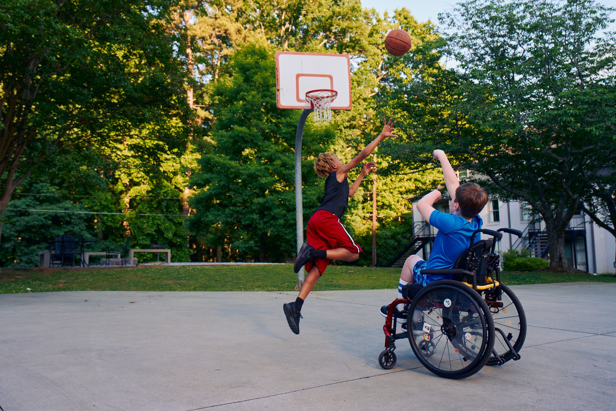 Two teens play basketball at a neighborhood park. One teen, a white boy seated in a manual wheelchair, shoots the ball. The other teen, a light-skinned African American male, jumps forward with arms outstretched, defending the shot. The teen seated in a manual wheelchair has short brown hair and is wearing blue athletic wear. The other teen is wearing black and red athletic wear.